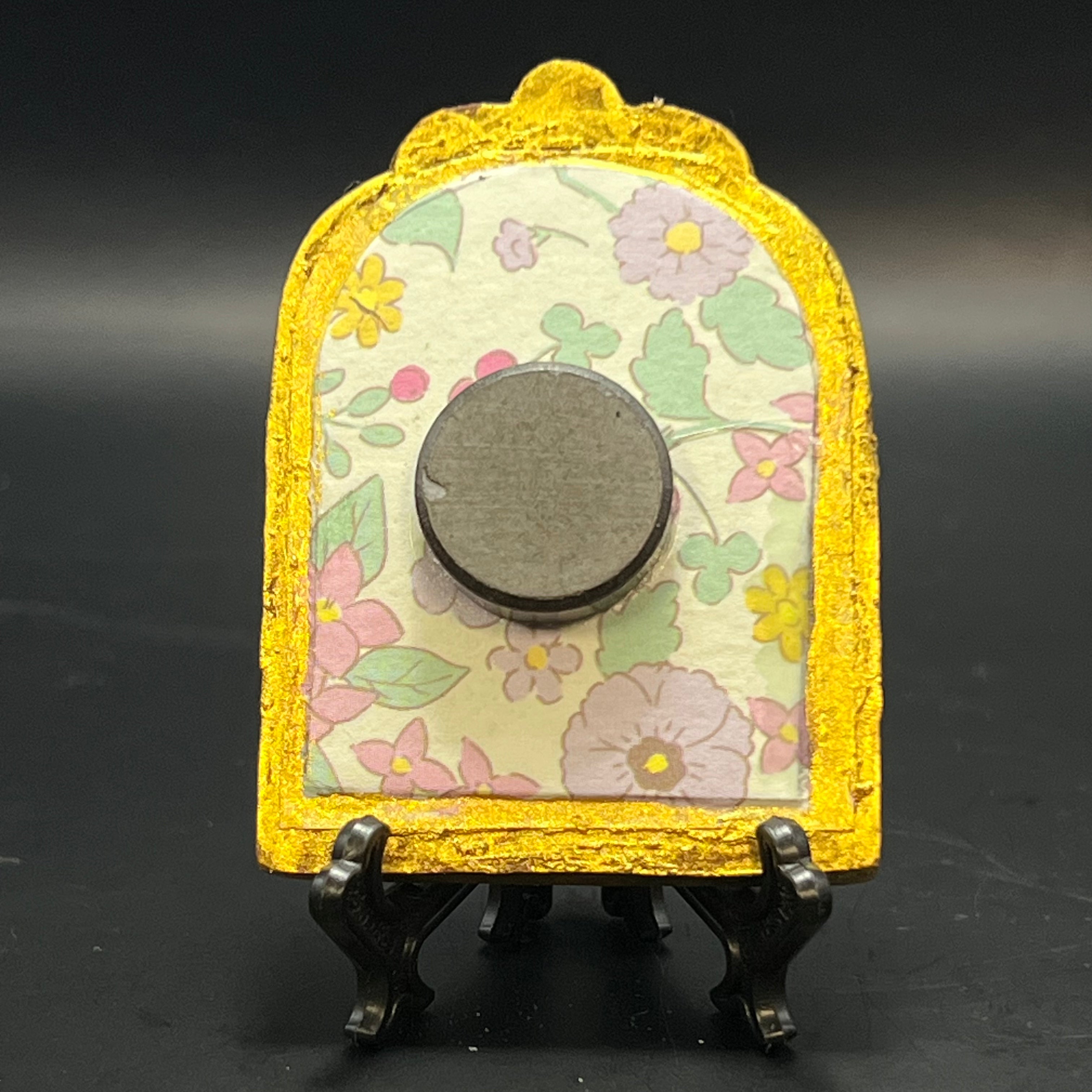 This is the Peonia Mini Painting by Chris Foster Design. This is showcasing the back. Backed with light green, pink, purple, and yellow floral pattern. It is against a dark background.