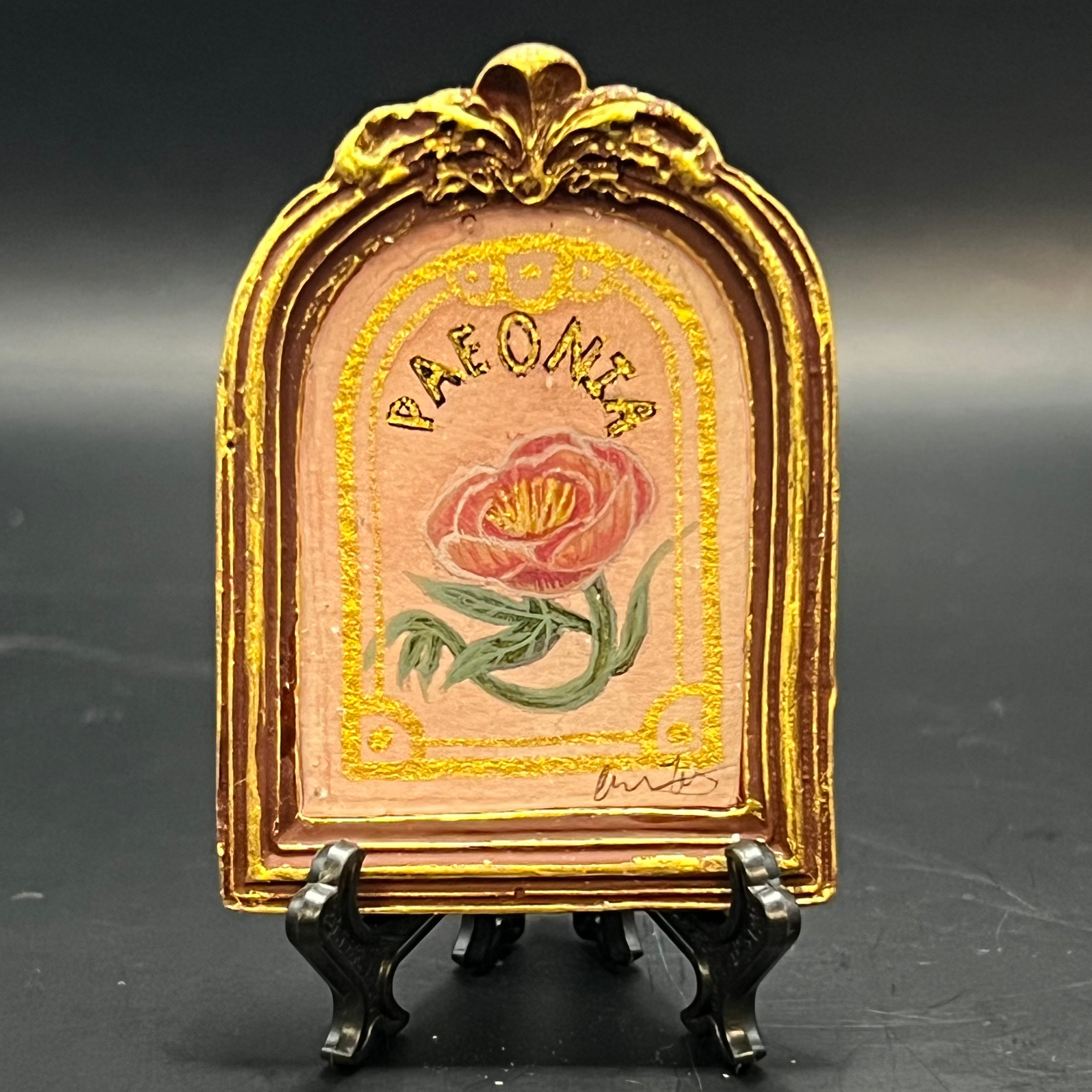 This is the Peonia Mini Painting by Chris Foster Design. This is showcasing the front. Ornate brown round top frame with gold distress. Coral charm peony on a light pink and gold glitter wash background with decorative gold border. It is against a dark background.
