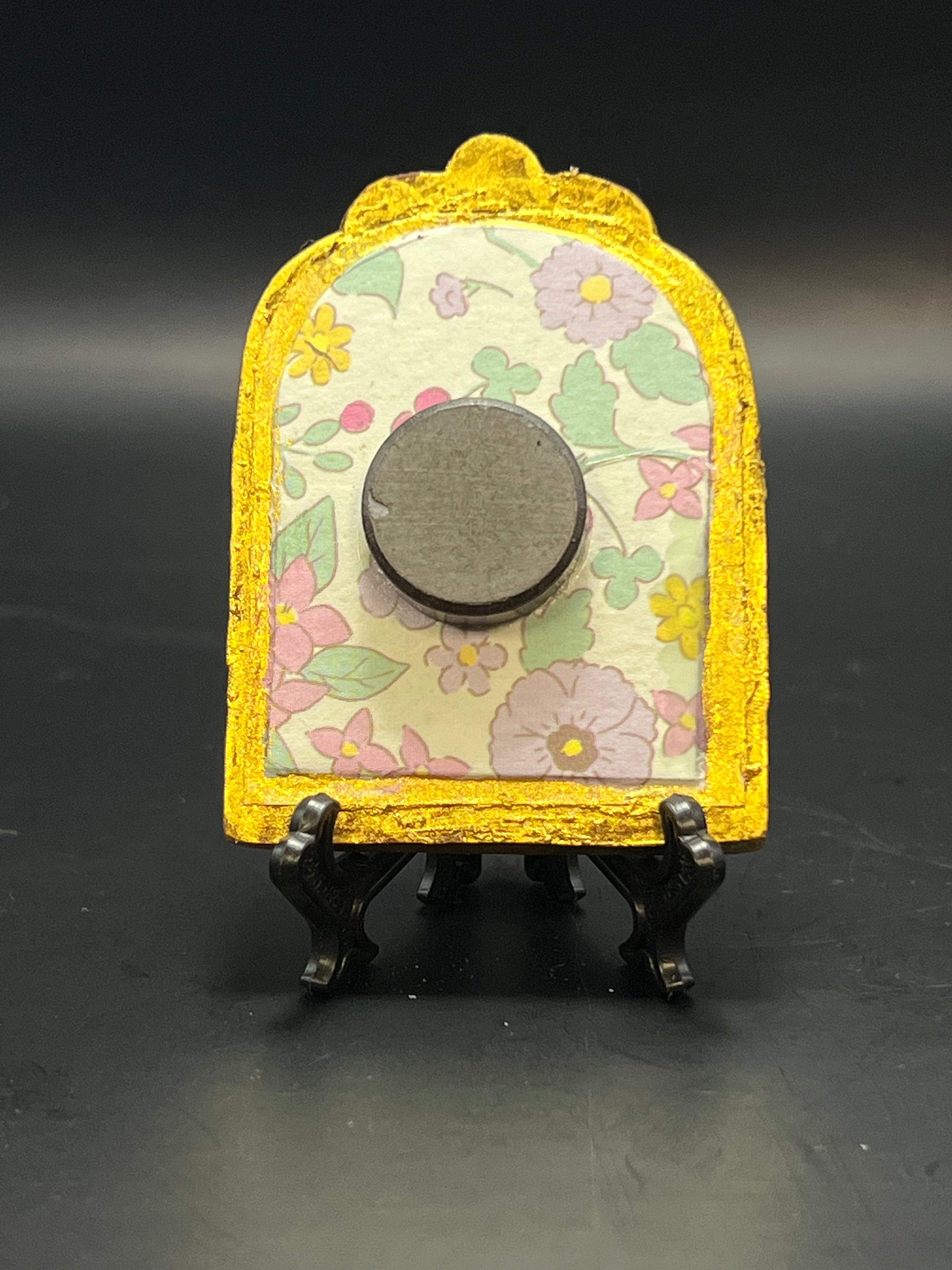 This is the Peony Mini Painting by Chris Foster Design. This showcases the back of the painting. There is a decorative gold frame where the backing is a floral design with purple, yellow and pink flowers with green leaves. There is a round magnet and is on a stand. This is all against a black background.