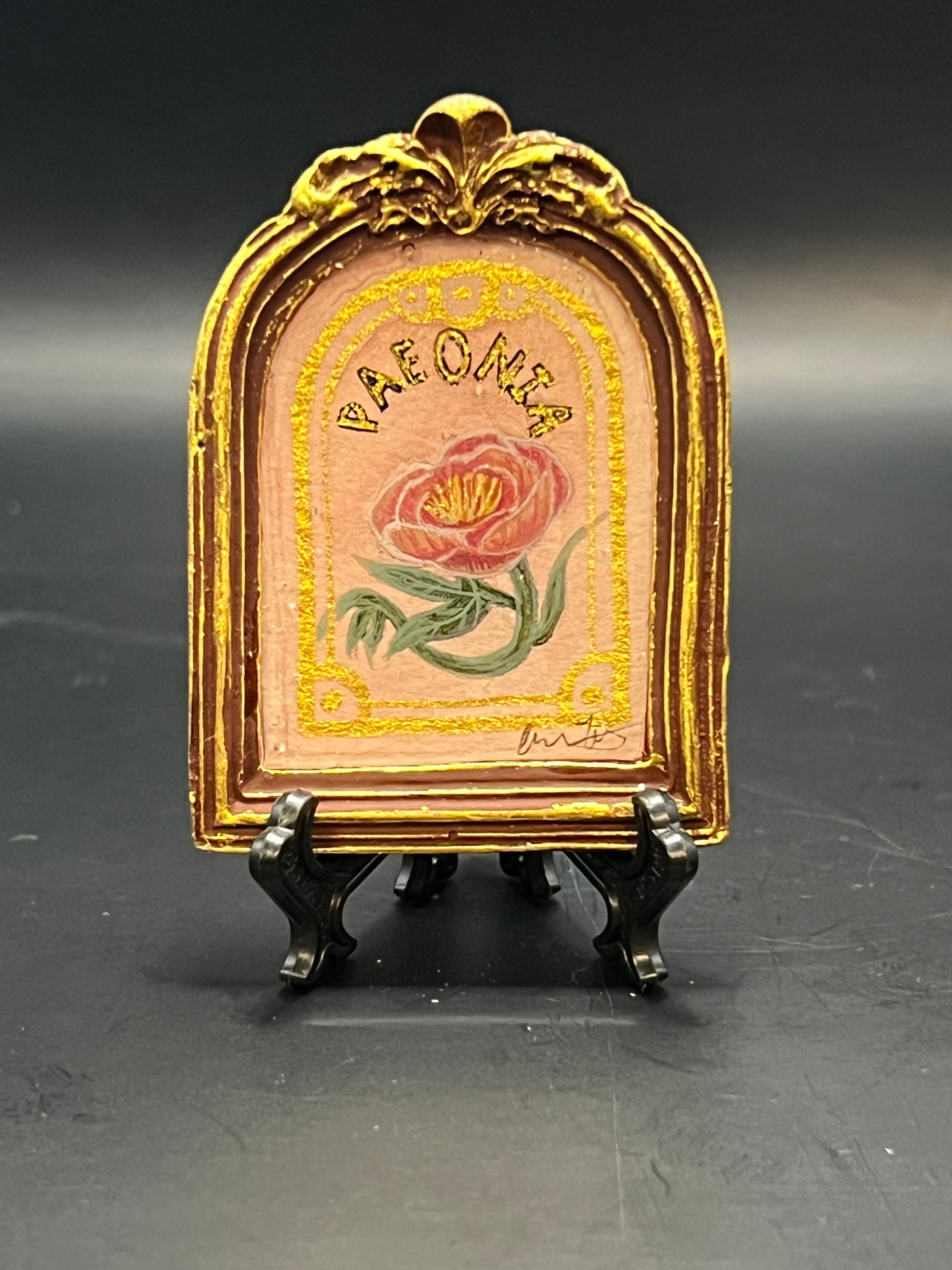 This is the Peony Mini Painting by Chris Foster Design. There is a gold decorative frame with a rose design and 'Paeonia' text in gold with a black outline. Around the rose and wording is a gold frame. The painting is against a light pink background. The studio's signature is on the bottom right. The painting frame is on a stand against a dark background.