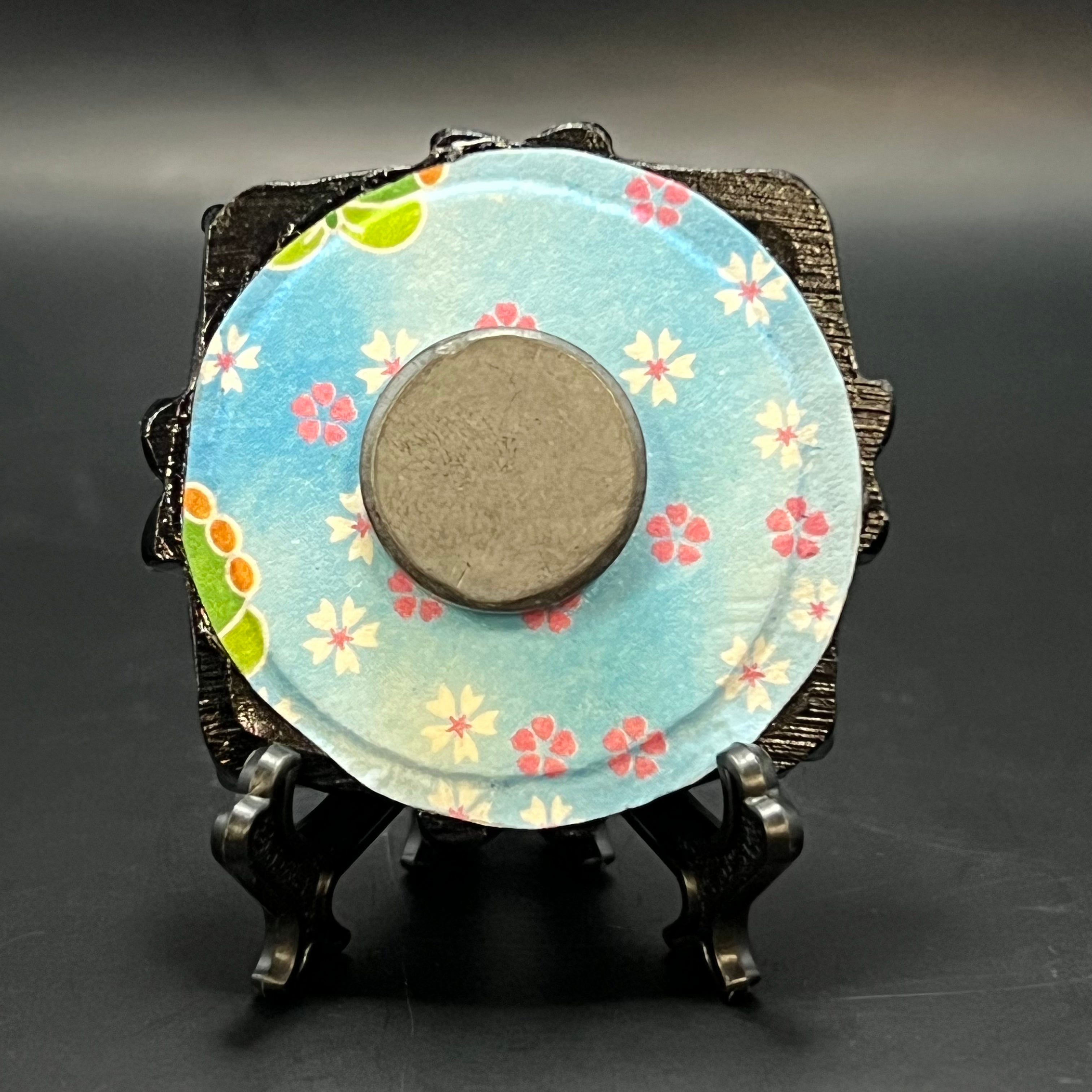 This is the Plum Blossom Bloom Circle by Chris Foster Design. This is showcasing the back. It is backed with blue, pink, and white floral origami paper. It is against a dark background.