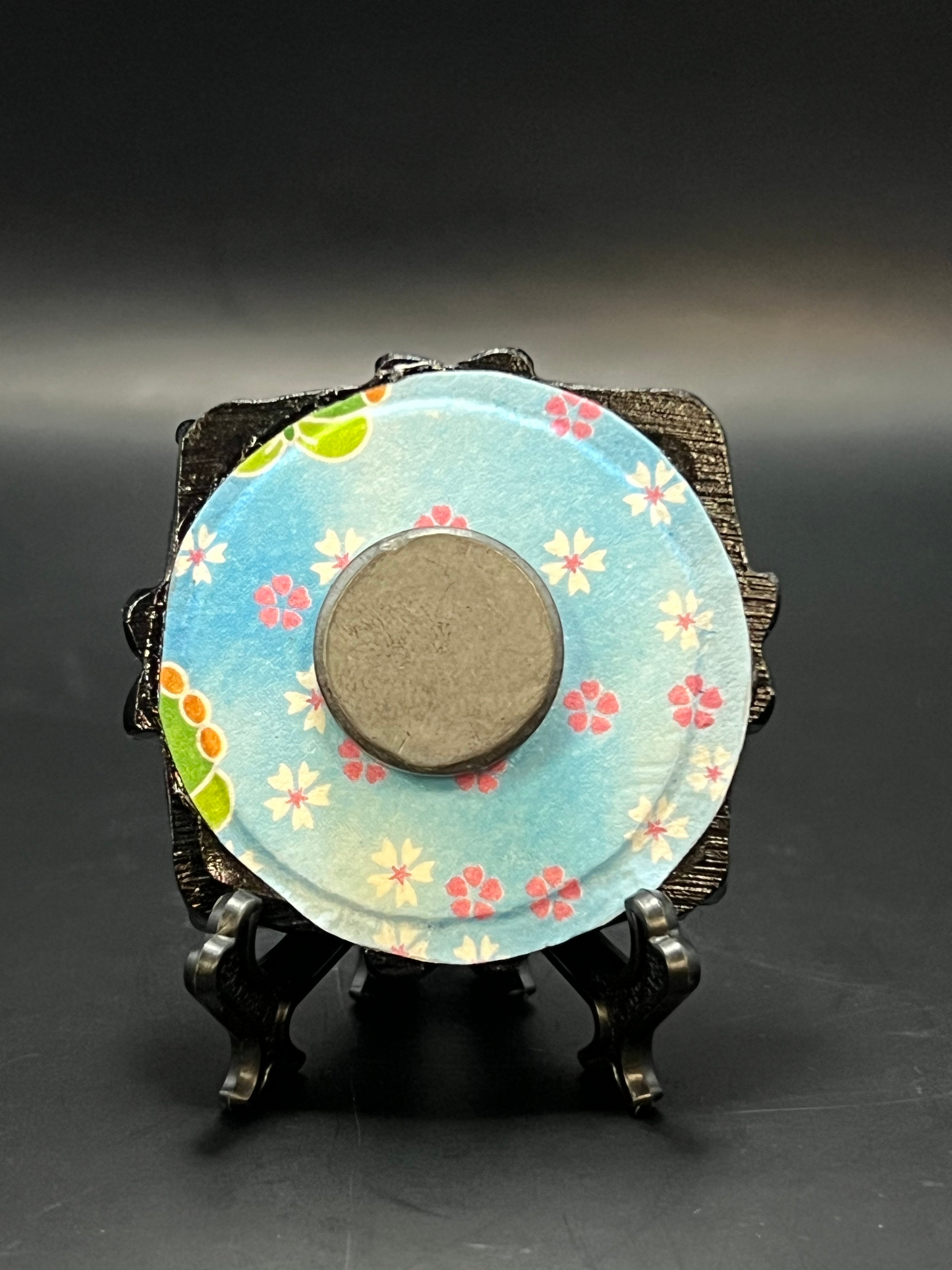 This is the Plum Blossom Bloom Circle Mini Painting by Chris Foster Design. There is a black decorative frame and a backing with floral patterns that are white and red with green leaves plus a light blue background. It is all on a stand and there is a round magnet against a dark background.