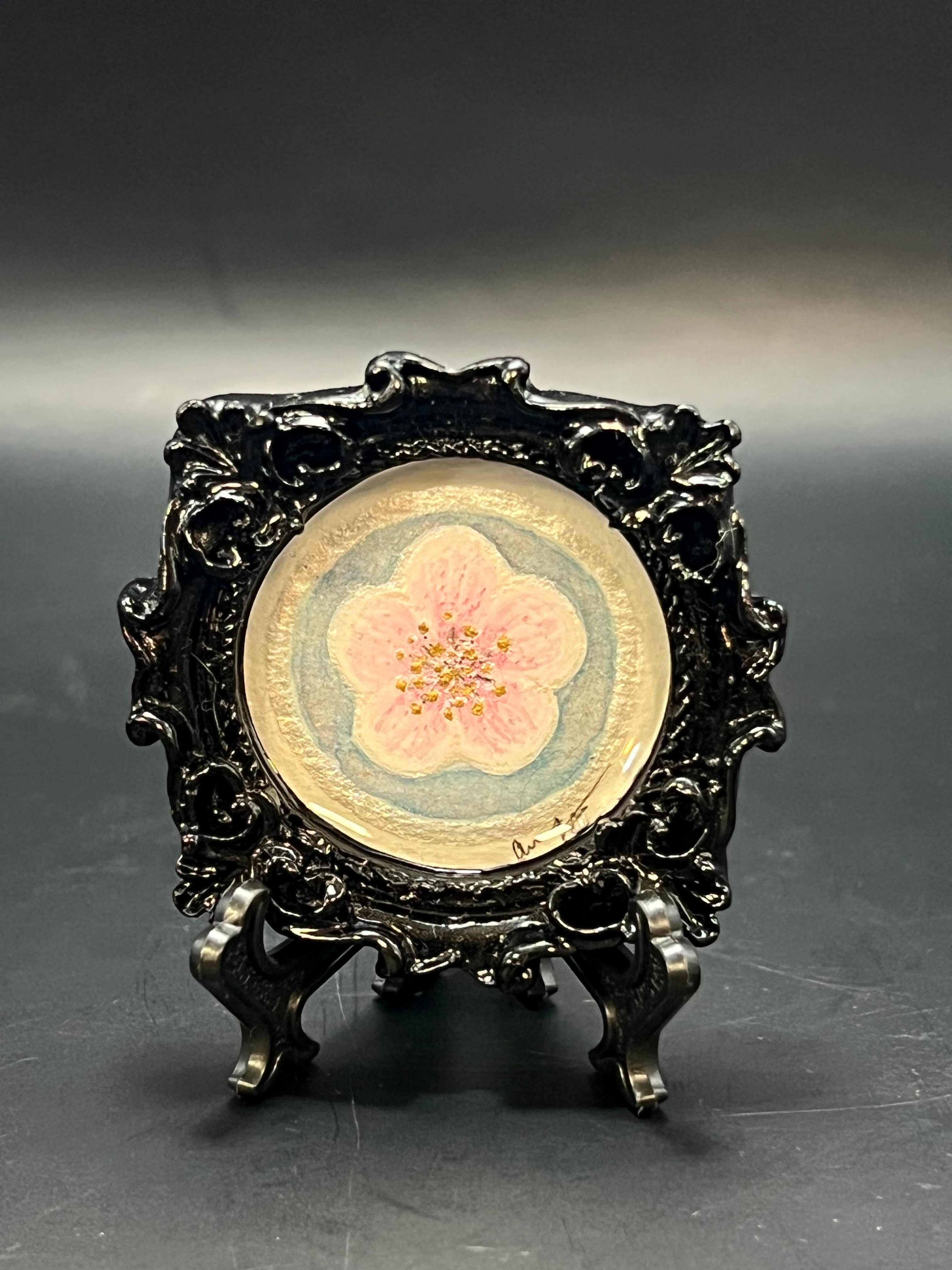 This is the Plum Blossom Bloom Circle by Chris Foster Design. This is showcasing the front of the painting. There is a black decorative frame with a pink flower design on a blue background and a white circle around everything on a dark gray background.