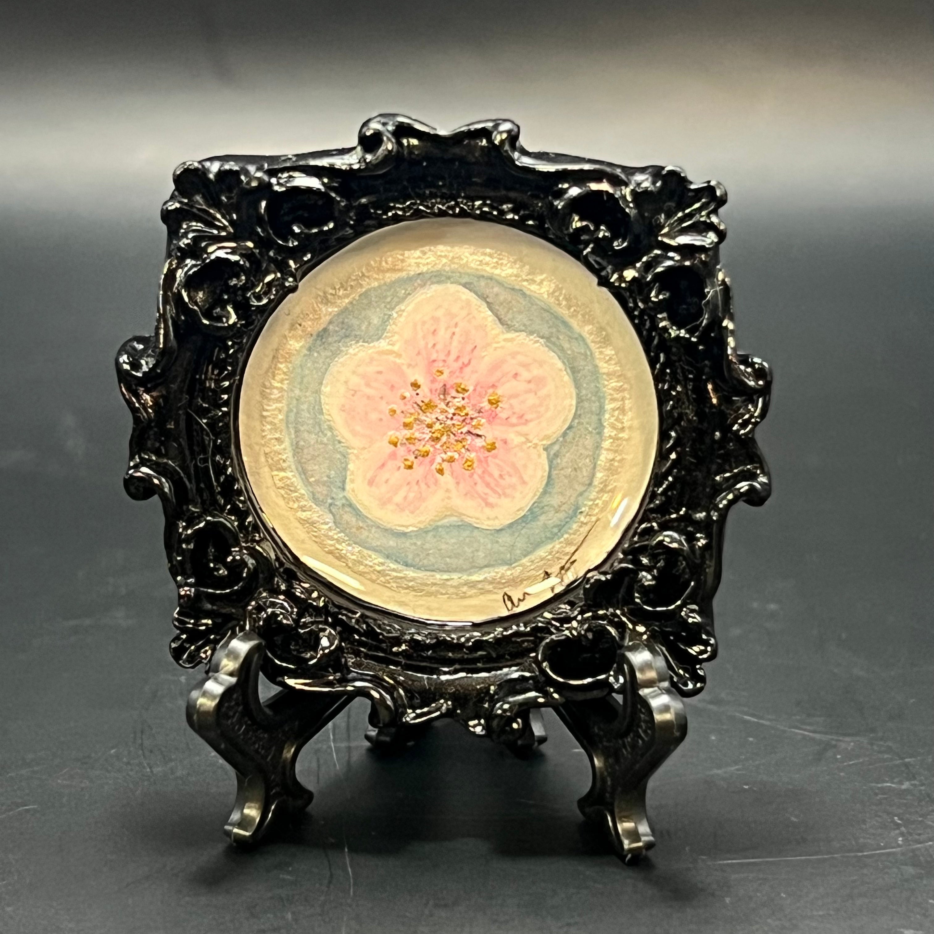 This is the Plum Blossom Bloom Circle Mini Painting by Chris Foster Design. This is showcasing the front. Black gloss ornate circle frame. Single light pink plum blossom bloom with silver outline and gold stamens on a light blue background with pink glitter wash. It is against a dark background.