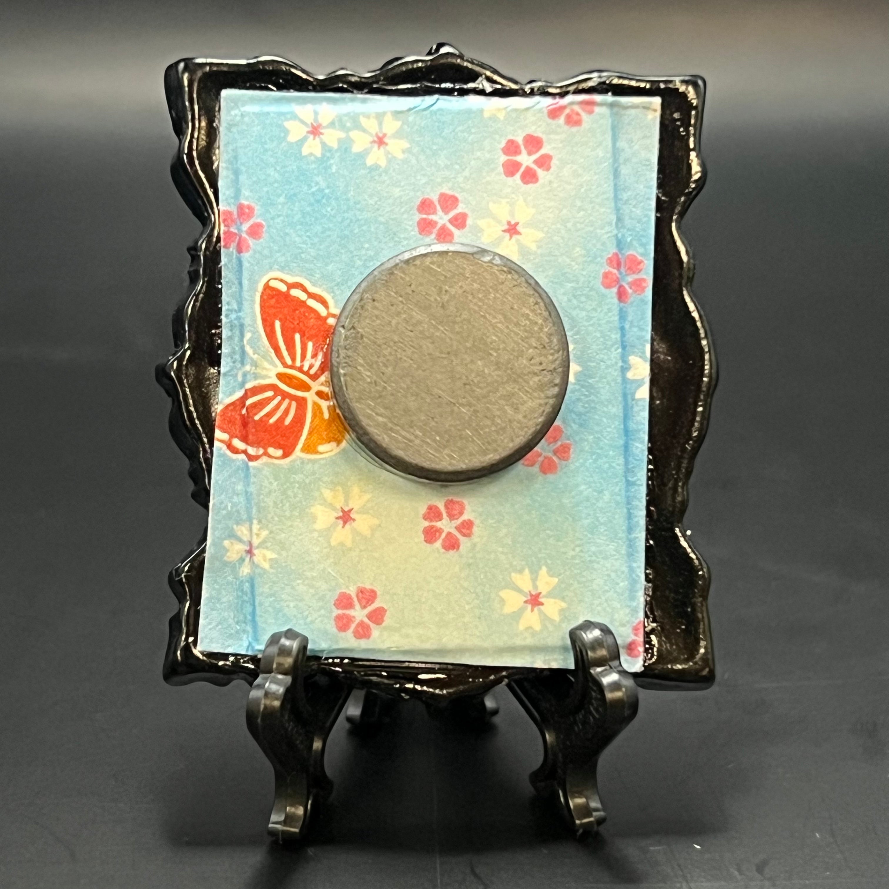 This is the Plum Blossom Rectangle Mini Painting by Chris Foster Design. This is showcasing the back. Backed with pink, white, red, and blue butterfly and floral pattern origami paper. It is against a dark background.