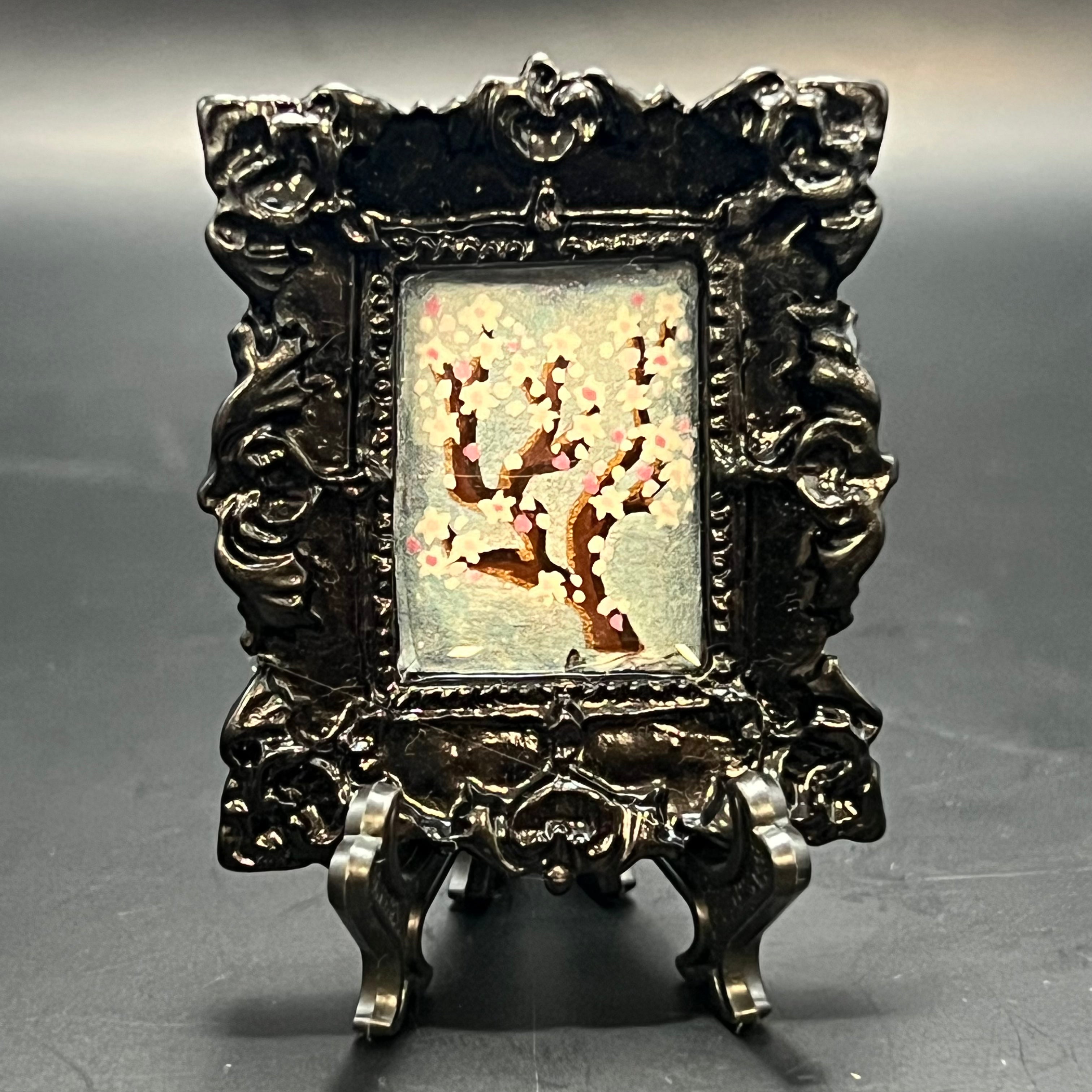 This is the Plum Blossom Square Mini Painting by Chris Foster Design. This is showcasing the front. Ornate rectangle gloss black frame. Plum blossom branch painting with light pink and white flowers on a light blue and pink glitter wash background. It is against a dark background.