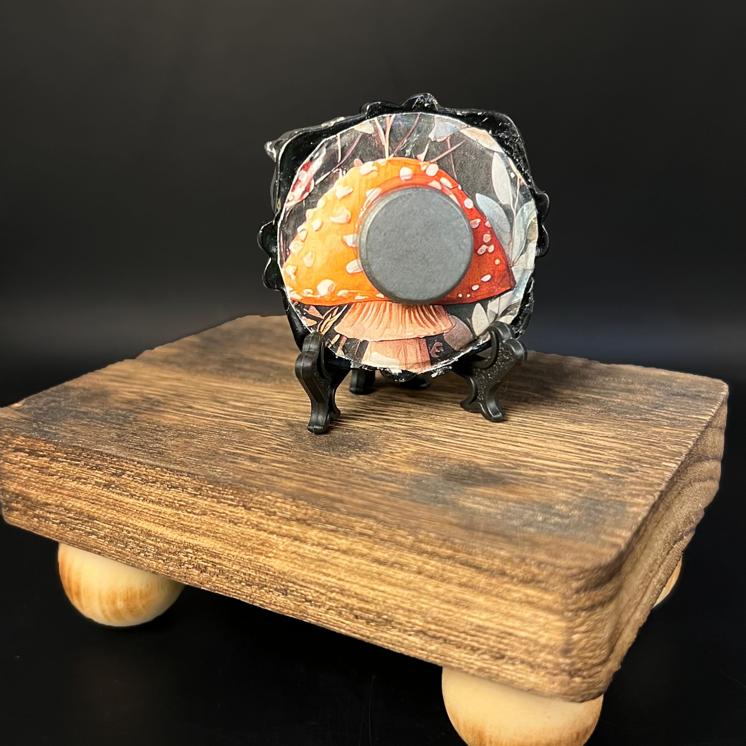 This is the Poison Mini Painting by Chris Foster Design. This is showcasing the back of the painting. There is a black circular frame and backed with a yellow mushroom, white flowers and black background design. The painting is on a wooden stand against a black background.