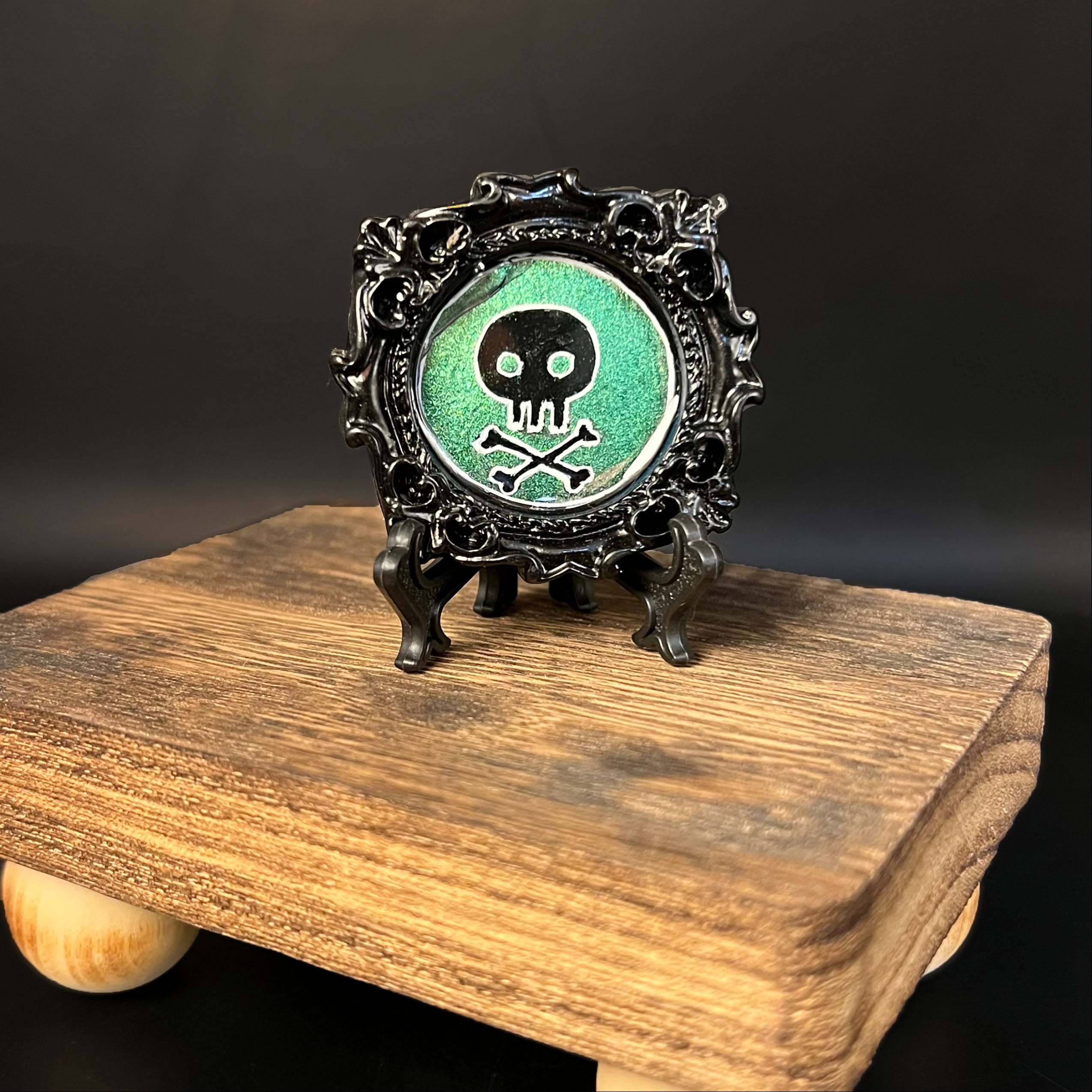 This is the Poison Mini Painting by Chris Foster Design. This is showcasing the front of the painting. It has a decorative circular black frame, on a black stand for the painting with a black and outlined white skull design painting. The backing is a glittery green and is on a wooden stand against a dark background.