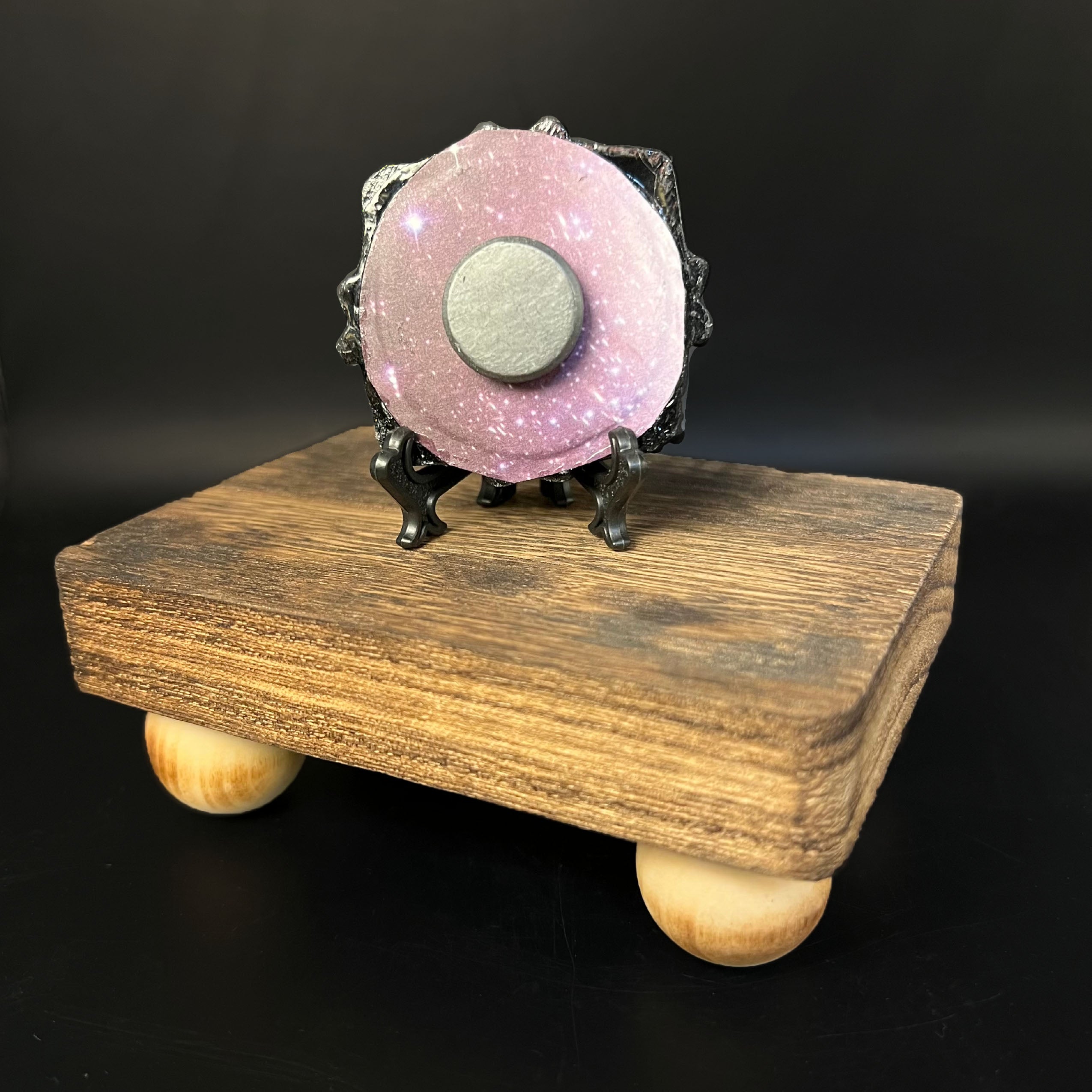 This is the Potion Mini Painting by Chris Foster Design. This is showcasing the back of the painting. The painting has a black circular frame and is backed with a pink, purple and black background with white/purple glistening stars. There's a round magnet in the middle with a black stand. The painting all together is on a wooden base against a black background.