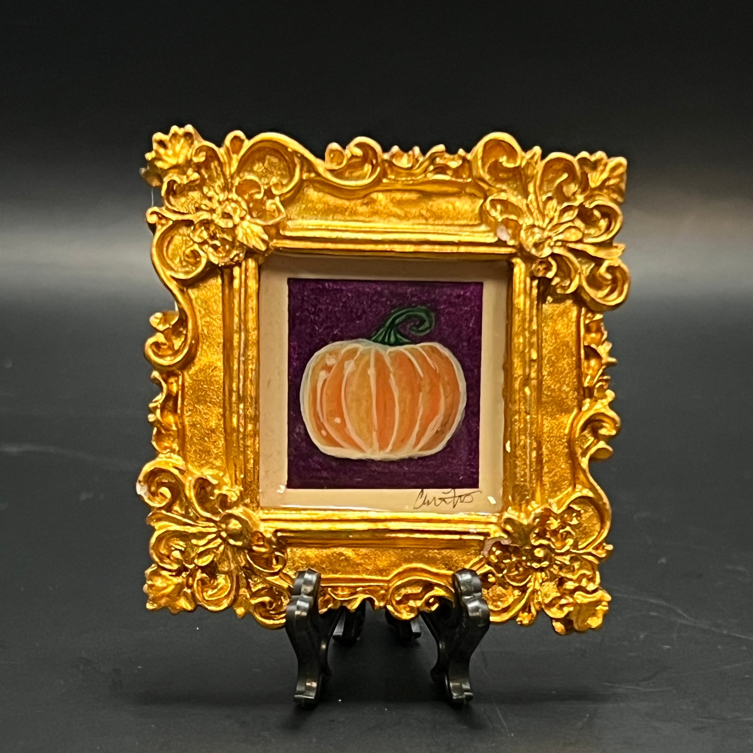 This is the Pumpkin Mini Painting by Chris Foster Design. Metallic Gold frame. Orange and white pumpkin painting on a glittered purple background with the studio's signature on the right. The painting is against a stand and dark background. 