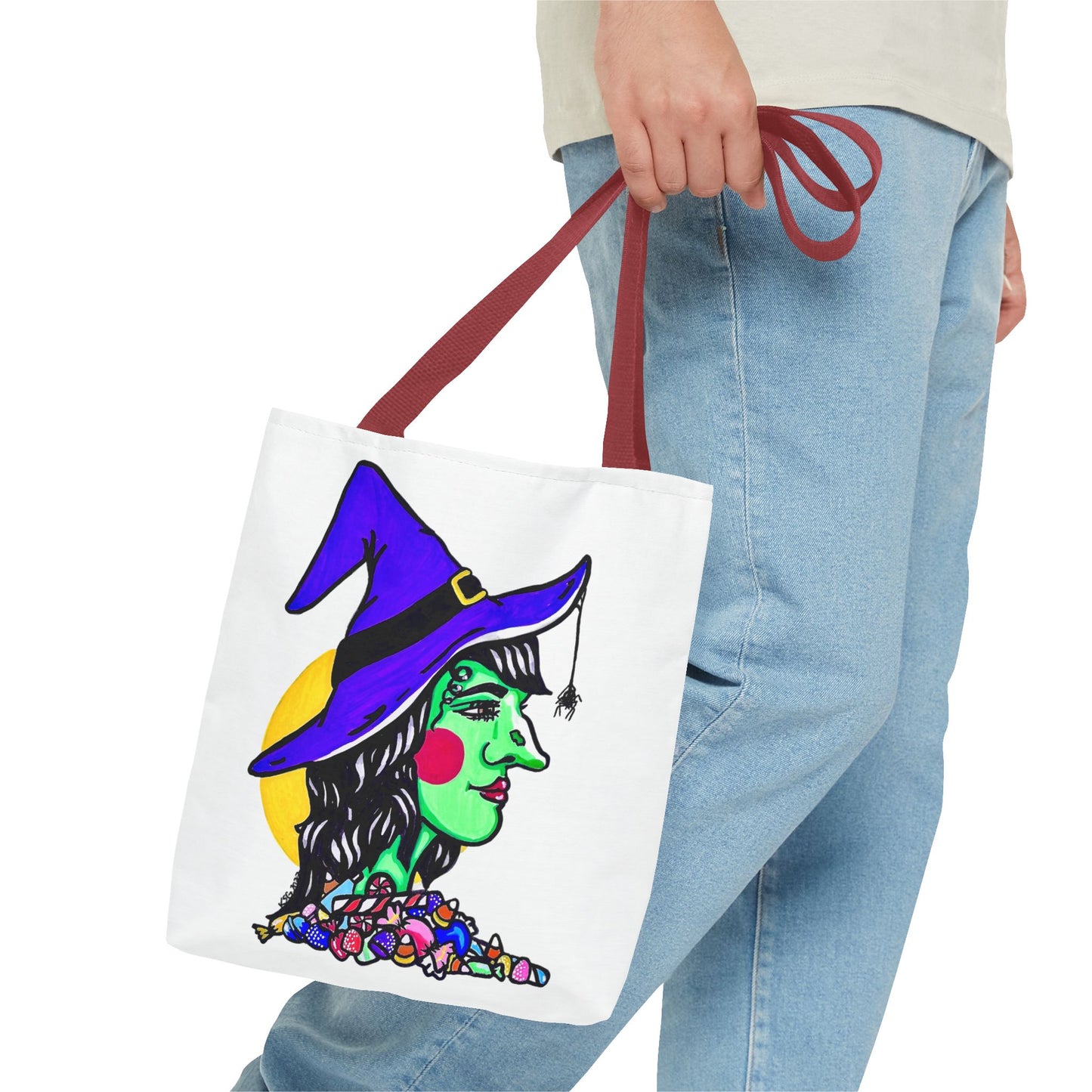 This is the Red 13" x 13" Witch Tote Bag by Storm Garden Studio. The handles are color matched. It shows the side profile of a witch. She has a purple witch hat, a black belt on hat, yellow buckle, a spider crawling down the front, red rosy cheeks, red lips and assorted candy under the neck. Behind her is a bright yellow circle past her hair. A man's hand with a beige tee and light- colored jeans is grabbing the handles from the hip down. The tote is against a white background.
