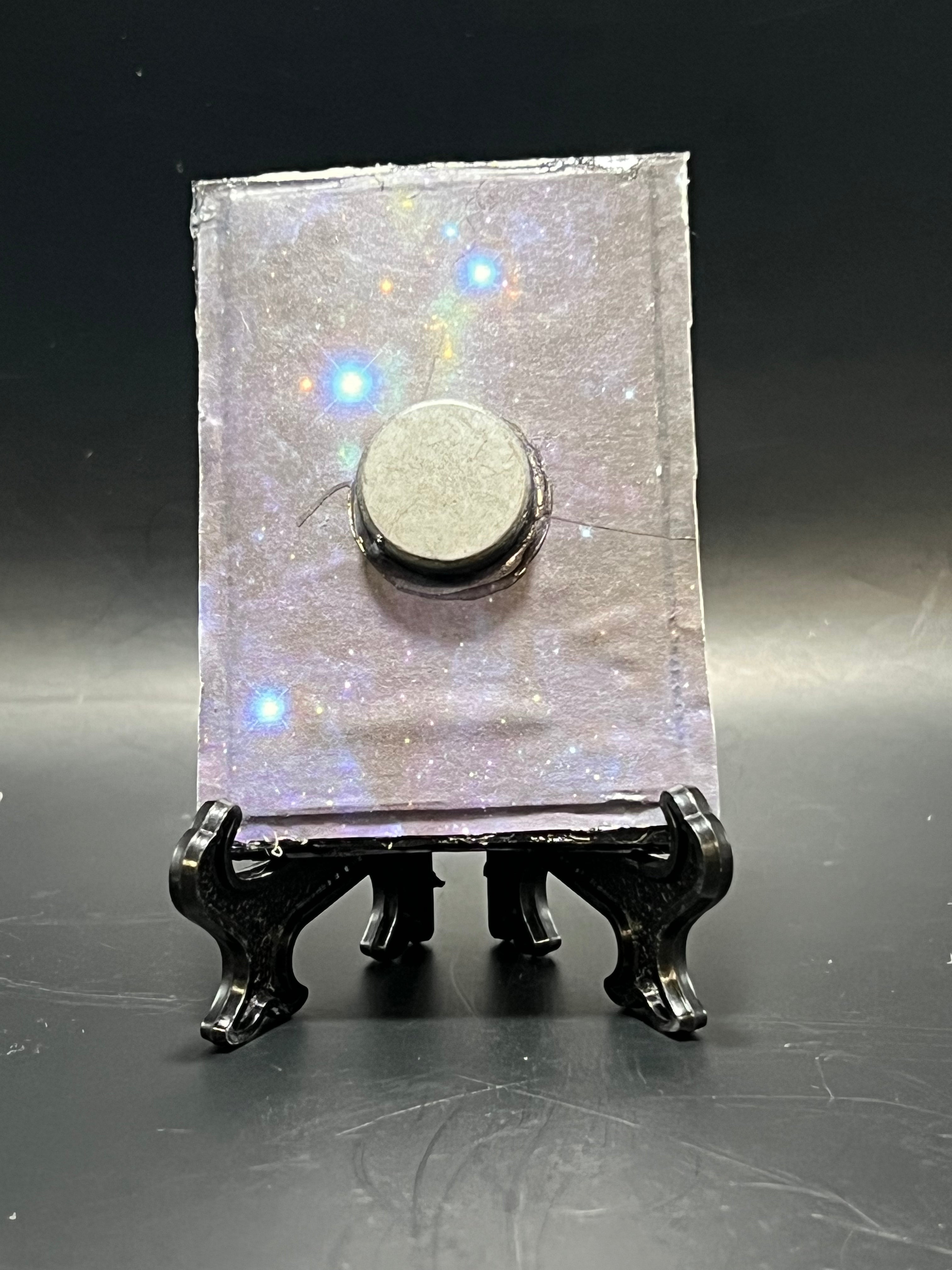 This is the handmade Saturn in Retrograde Miniature Painting by Chris Foster Design. This is showcasing the back. This is a small rectangular frame and the decorative item with a sparkly mix background with blues and blacks on a stand with yellow and blue stars. There is a magnet and against a miniature picture frame.