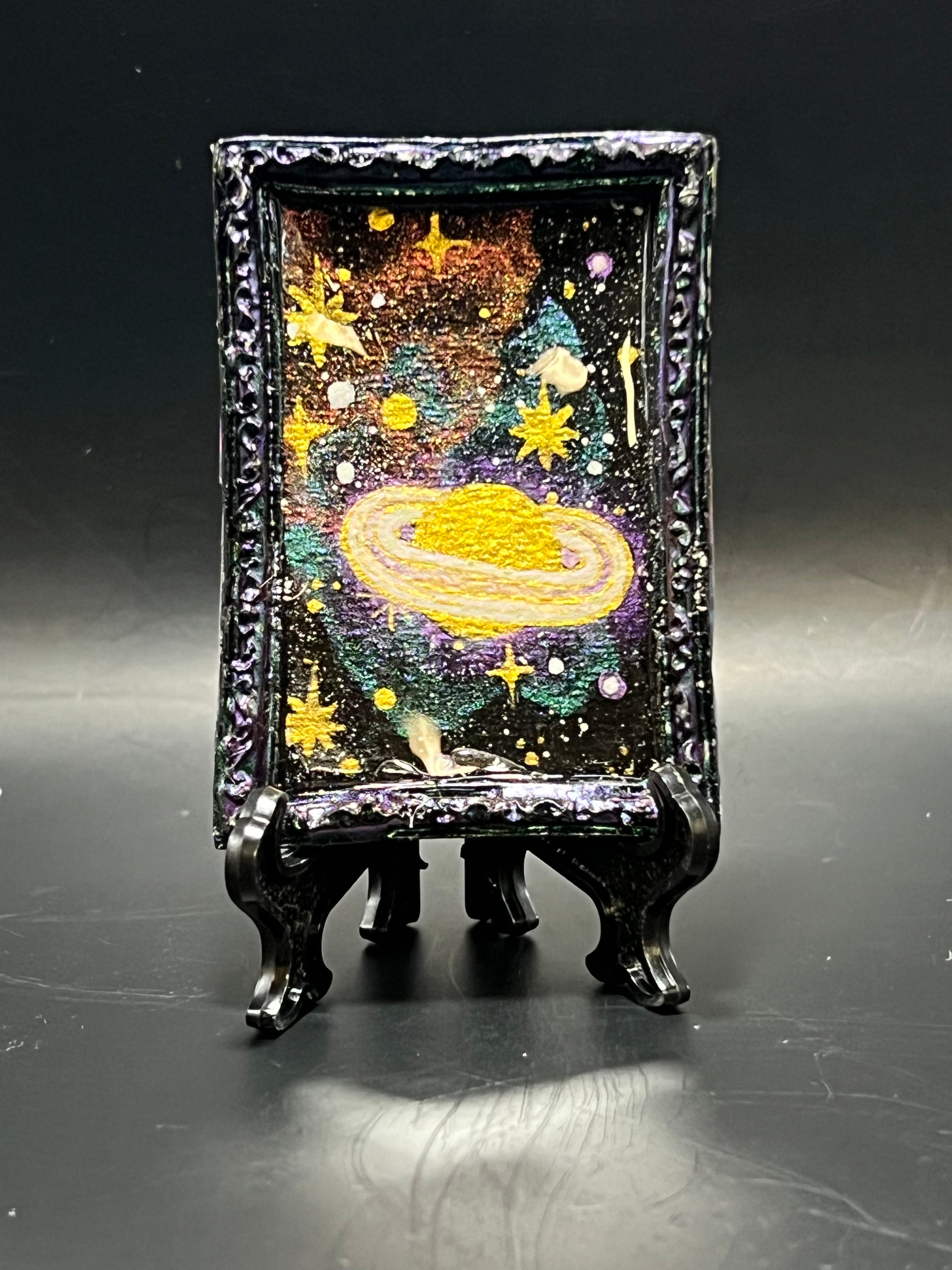 This is the handmade Saturn in Retrograde Miniature Painting by Chris Foster Design. This is showcasing the front. This is a decorative piece with a shimmery black frame, starry night design with Saturn to the right including larger yellow stars, sparkling green, orange, yellow and white smaller stars on a dark background. There is a magnet and against a miniature picture frame.