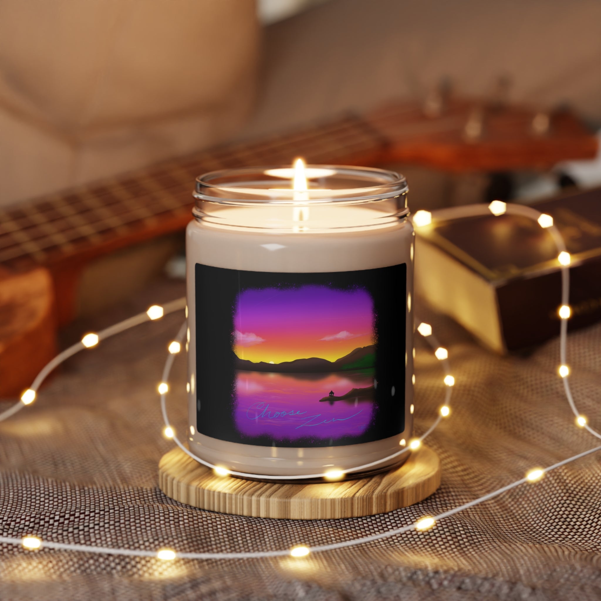 This is the Sea Salt+Orchid Choose Zen Scented Soy Candle by Lee Hansheng Studios. The illustration is a landscape when the sun is setting from a lake.It's surrounded by black like an old time photo and phases into purple,pink,red,orange then yellow where you can see the darker green hills.The candle has a 'Choose Zen' illustration on the label, placed on a wooden coaster, surrounded by a string of LED lights and a guitar in the background.