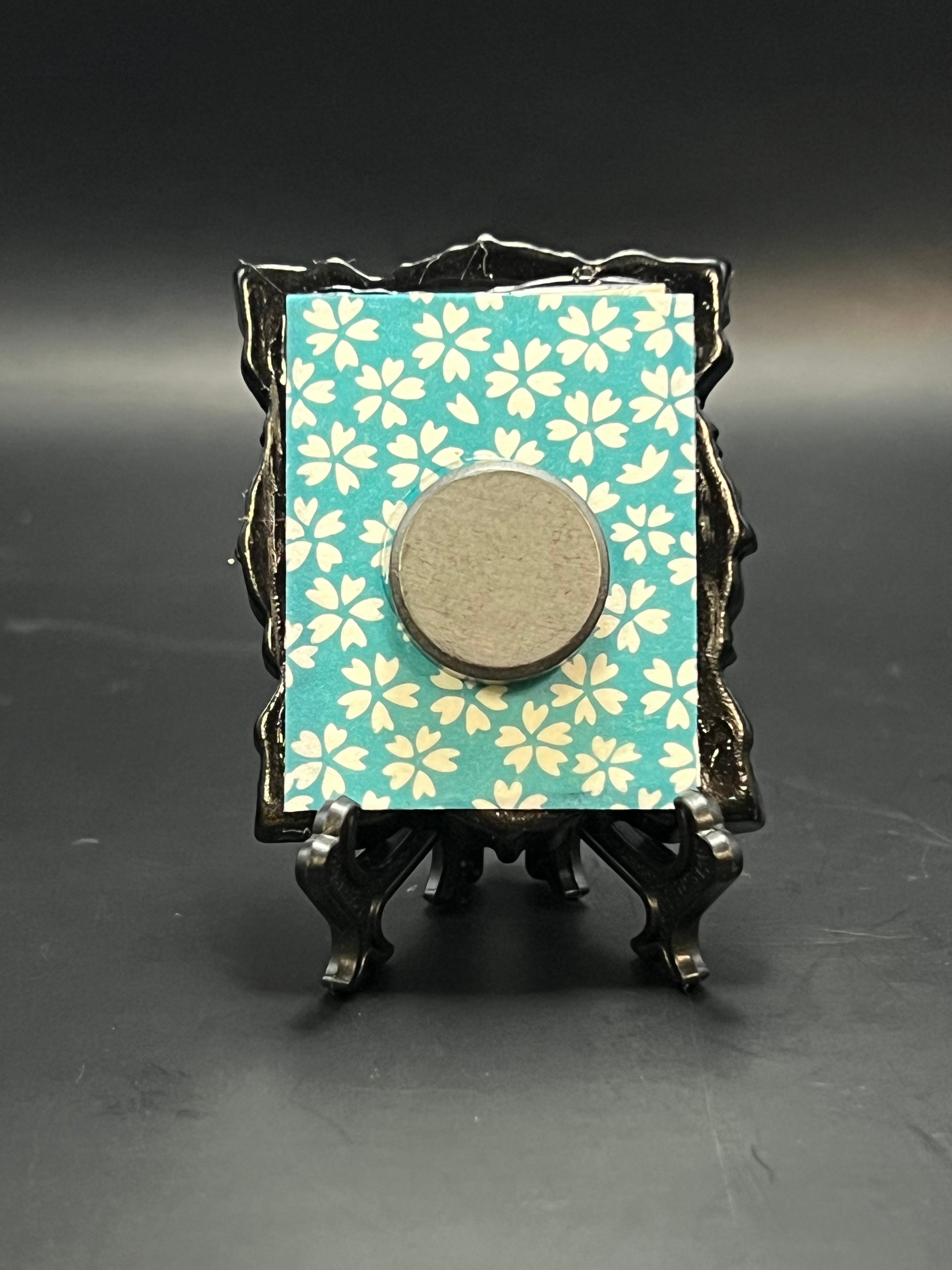 This is the Small Beetle Mini Painting by Chris Foster Design. This is showcasing the back of the painting. It is on a small black decorative frame with a turquoise background featuring white floral patterns backing. There is a round magnet on the back and is against a dark background.