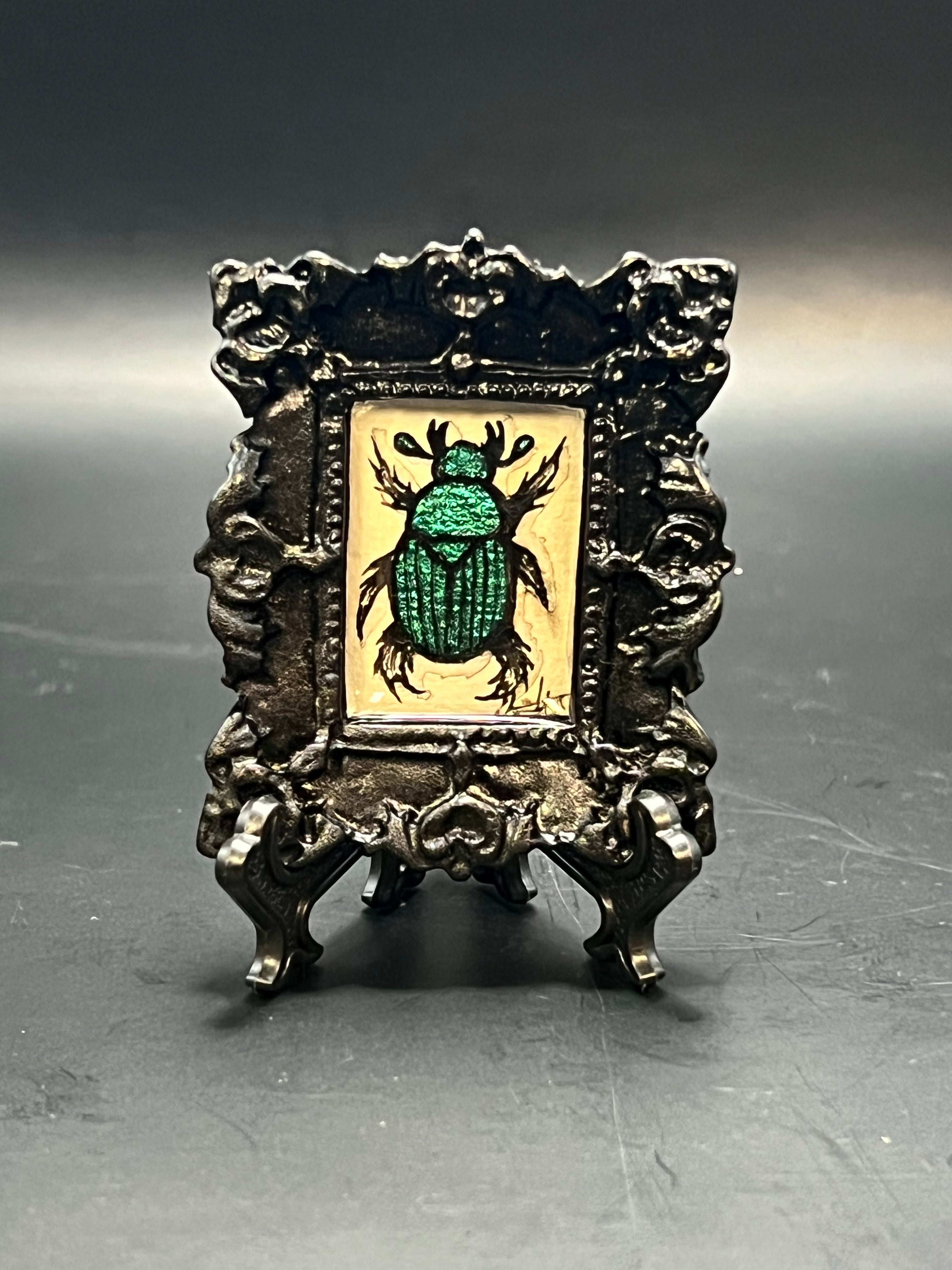 This is the Small Beetle Mini Painting by Chris Foster Design. This is showcasing the front of the painting. There is a black decorative frame with a glimmering big green beetle illustration on a tan/yellow background. The painting with the frame and stand is against a dark background. 