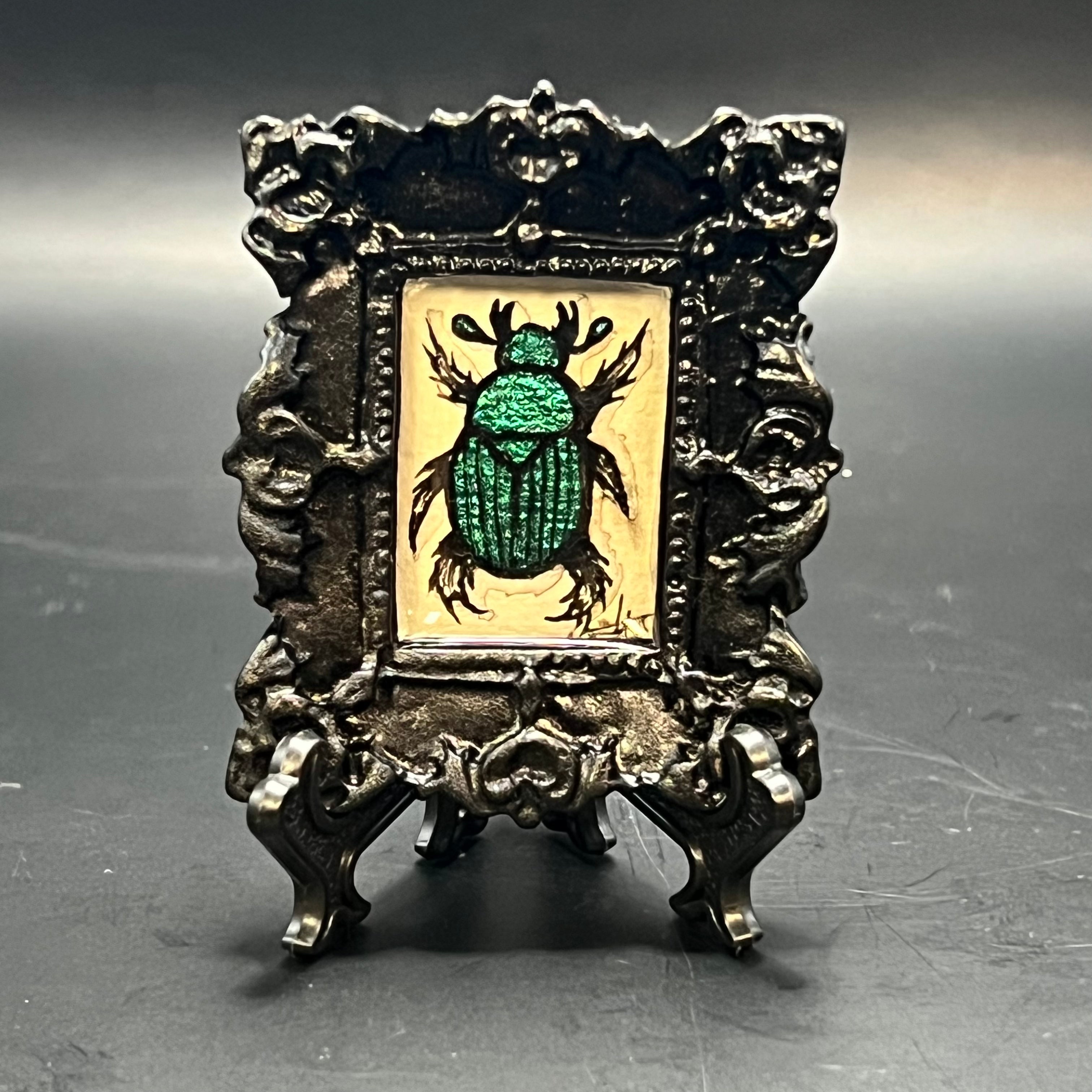 This is the Small Beetle Mini Painting by Chris Foster Design. This is showcasing the front. Ornate gloss black ornate frame. Color shift purple, blue, and green beetle on beige background. It is against a dark background. 