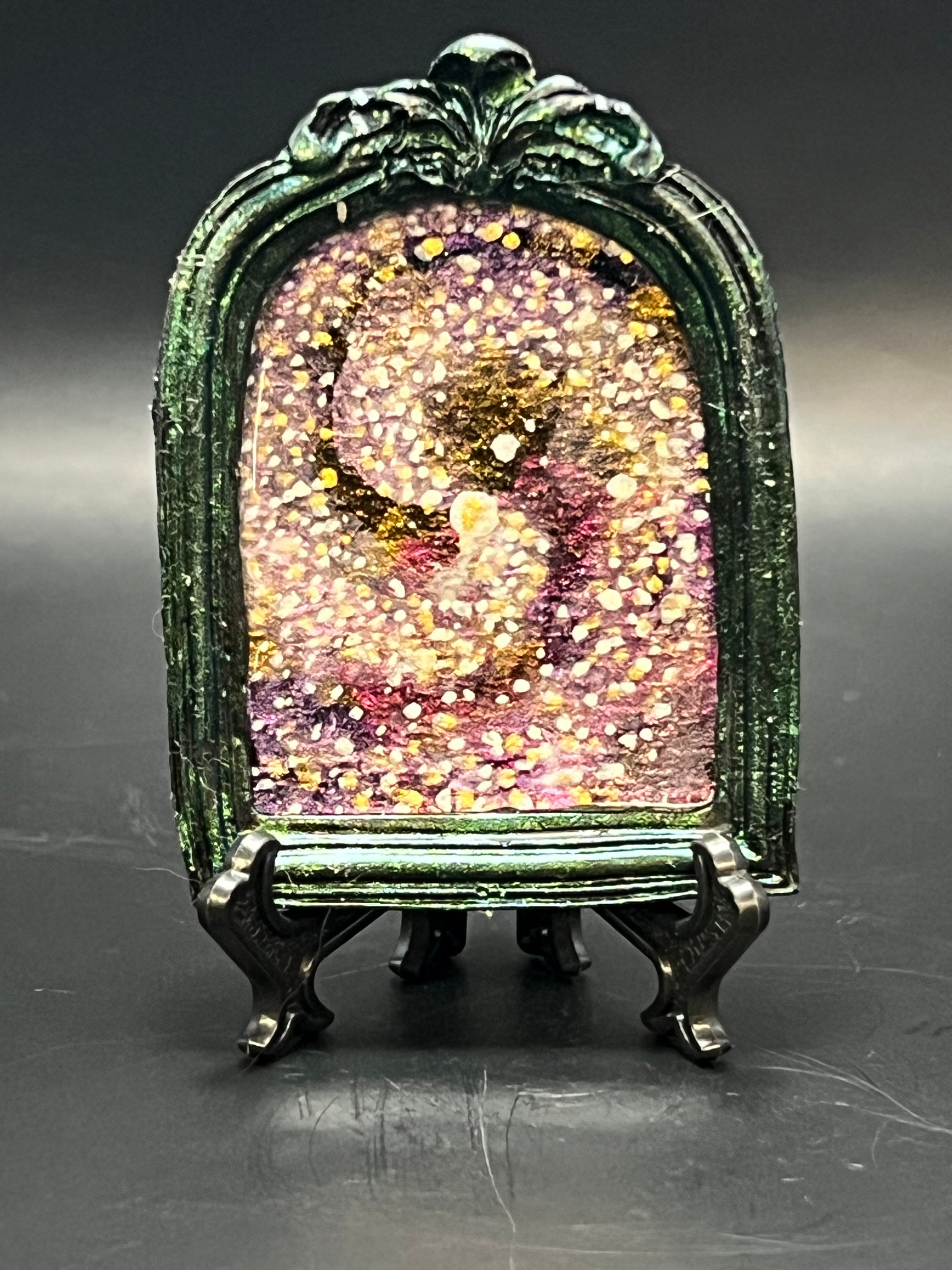 This is the handmade Spiral Galaxy Miniature Painting by Chris Foster Design. This is showcasing the front. Decorative piece with a colorful, galaxy-like design on a stand against a dark background. It is a decorative piece with a green frame and glittery center on a dark background. There is a magnet and against a miniature picture frame.