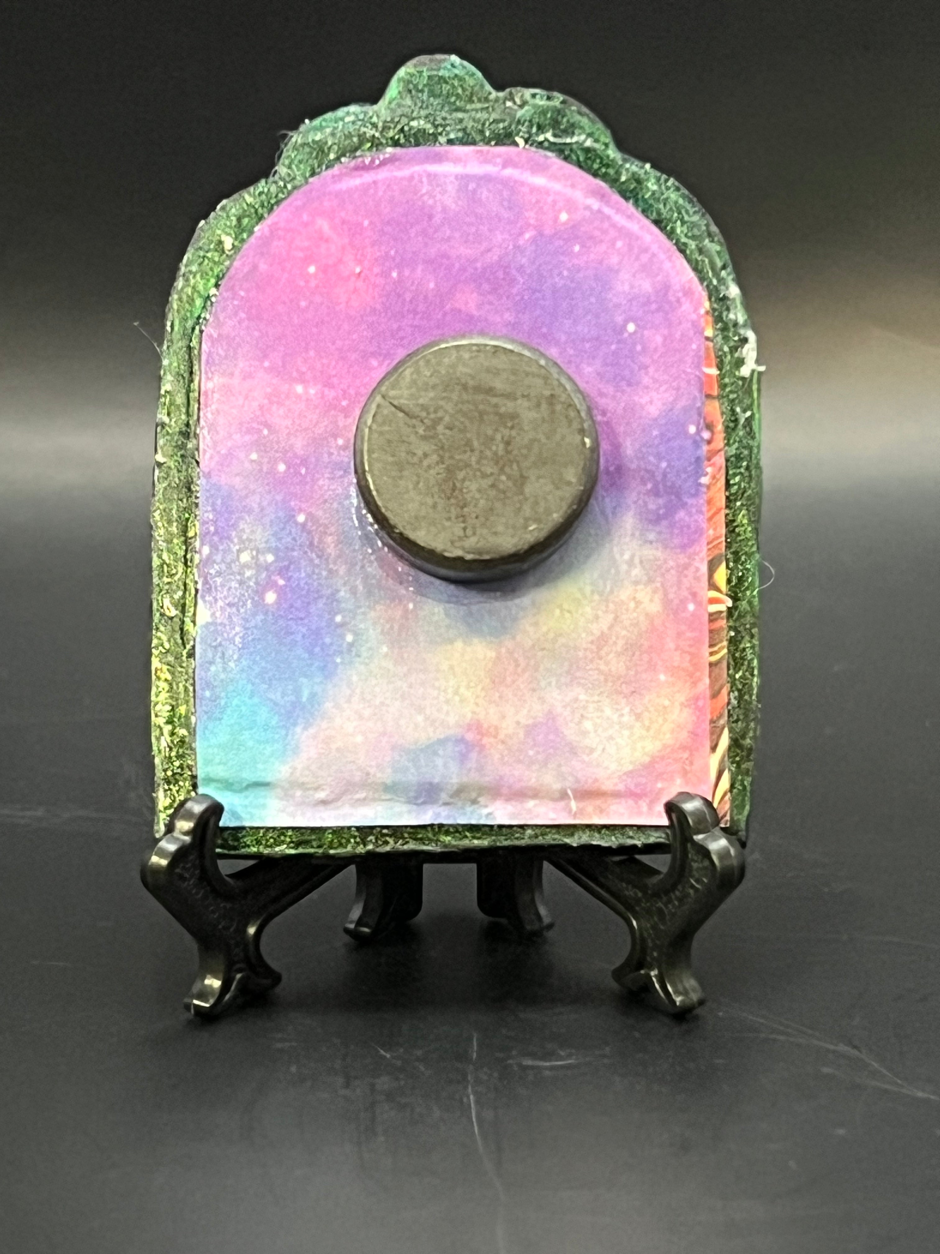 This is the handmade Spiral Galaxy Miniature Painting by Chris Foster Design. This is showcasing the back. Decorative piece with a colorful, galaxy-like design on a stand against a dark background. There is a magnet and against a miniature picture frame.
