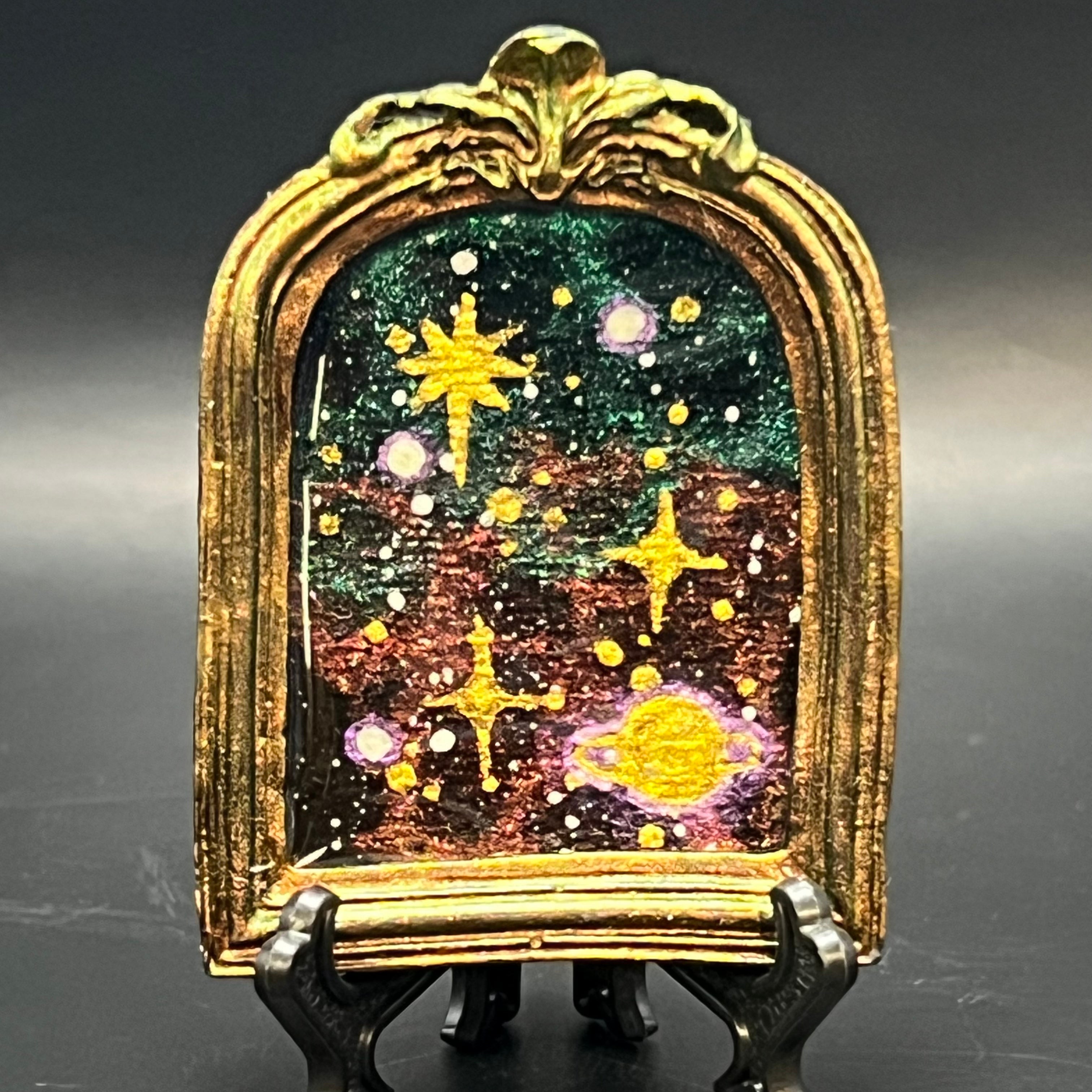 This is the Starry Orbit Mini Painting by Chris Foster Design. This is showcasing the front. There is an oval, shimmery gold decorative frame with starry night design, mix of green and orange in the background, twinkling yellow stars in different shapes, Saturn on the bottom right corner. The mini is on a black stand and against a dark background.