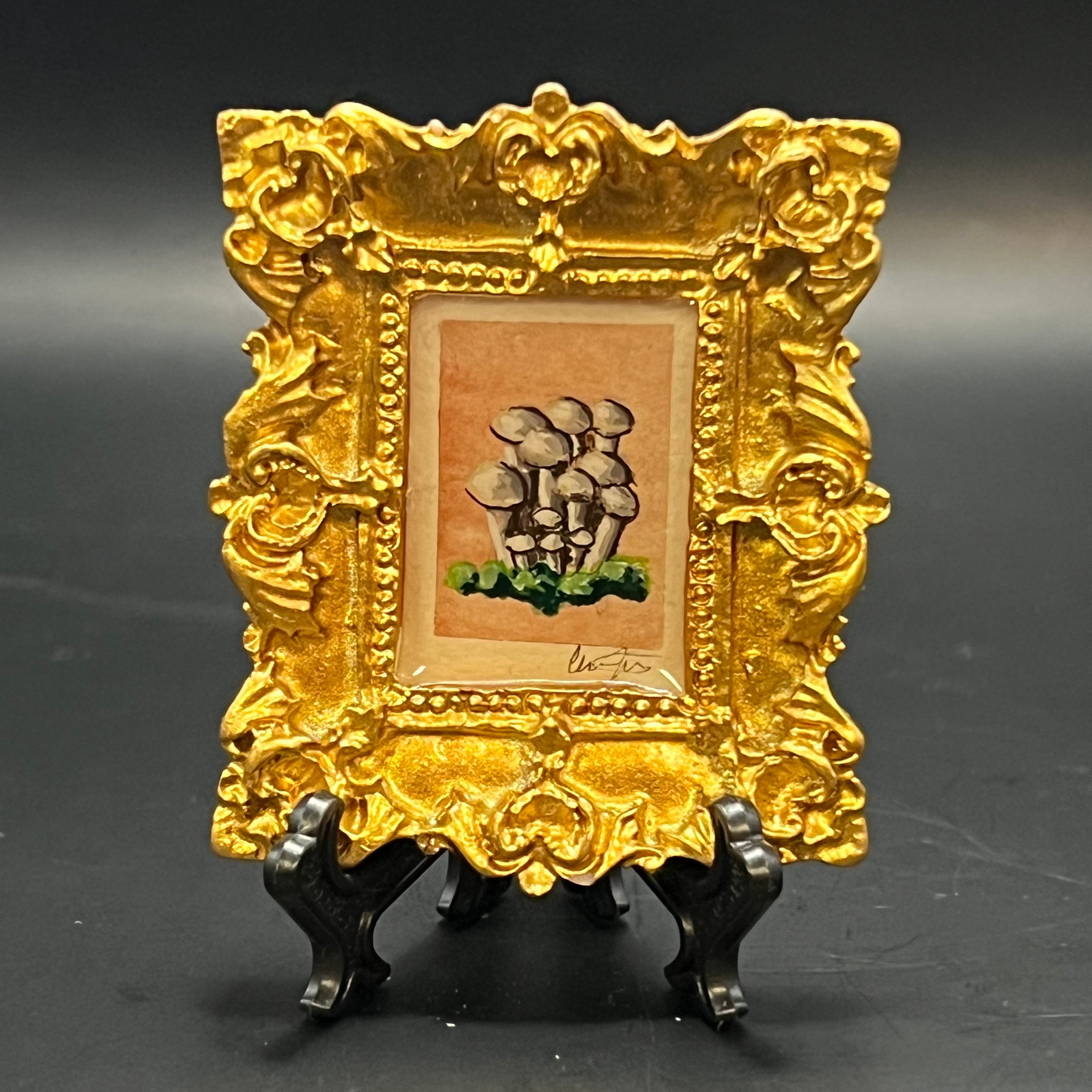 This is the Straw Mushroom Mini Painting by Chris Foster Design. This is showcasing the front. Ornamental metallic gold rectangular frame. Light grey straw mushrooms clustered amongst green moss on a peach watercolor background. It is against a dark background.