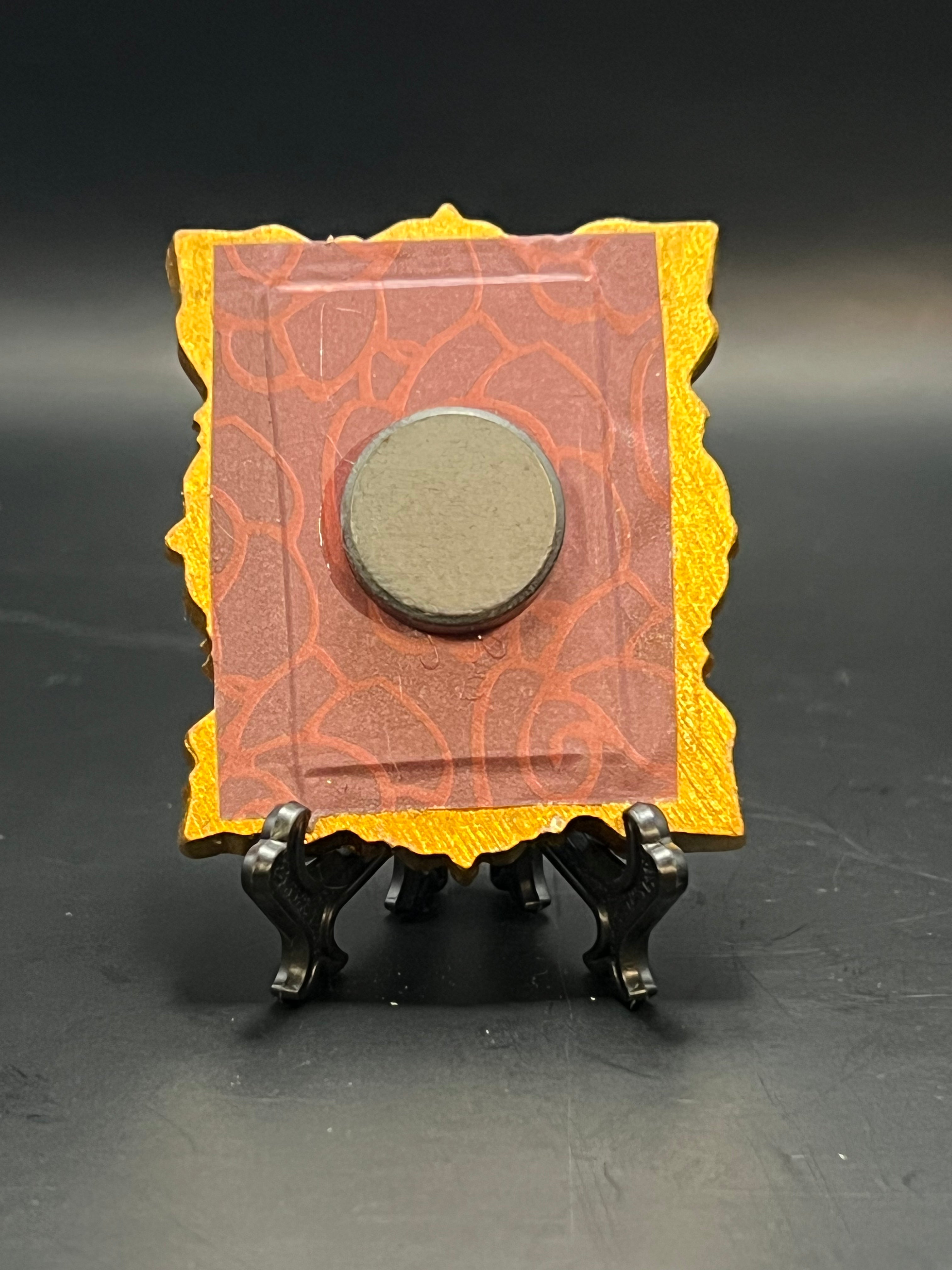 This is the handmade Straw Mushroom Miniature Painting by Chris Foster Design. This is showcasing the back. This is a small rectangular frame and the decorative item with a red and orange squiggly pattern background. There is a magnet and against a miniature picture frame.
