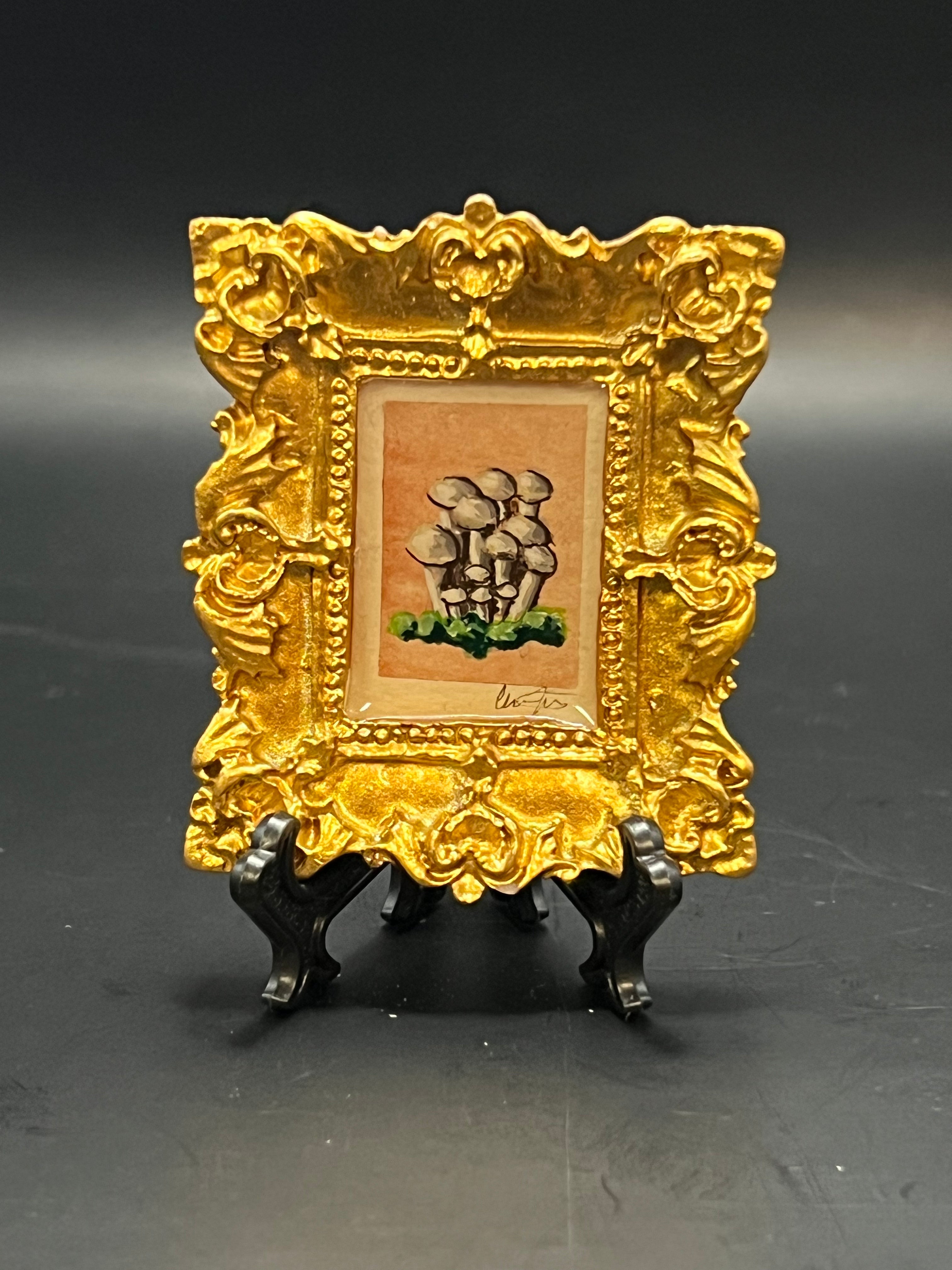 This is the handmade Straw Mushrooms Miniature Painting by Chris Foster Design. This is showcasing the front. This is a decorative piece with a gold rectangular frame, there are a bundle of mushrooms in different sizes that are gray on top of dark green grass. The background is light orange.
