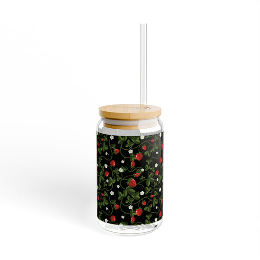 This is the 16 oz Strawberry Sipper Glass by Chris Foster Designs. The glass comes with a bamboo lid and Tritan plastic straw. The image is from the right with straw facing the same way. The glass has a wrap around illustration that has been printed. It is an image of red strawberries, white growing flowers with lush green leaves and green vining patterns that are throughout. The background on the image is black. The sipper glass is against a white background. 