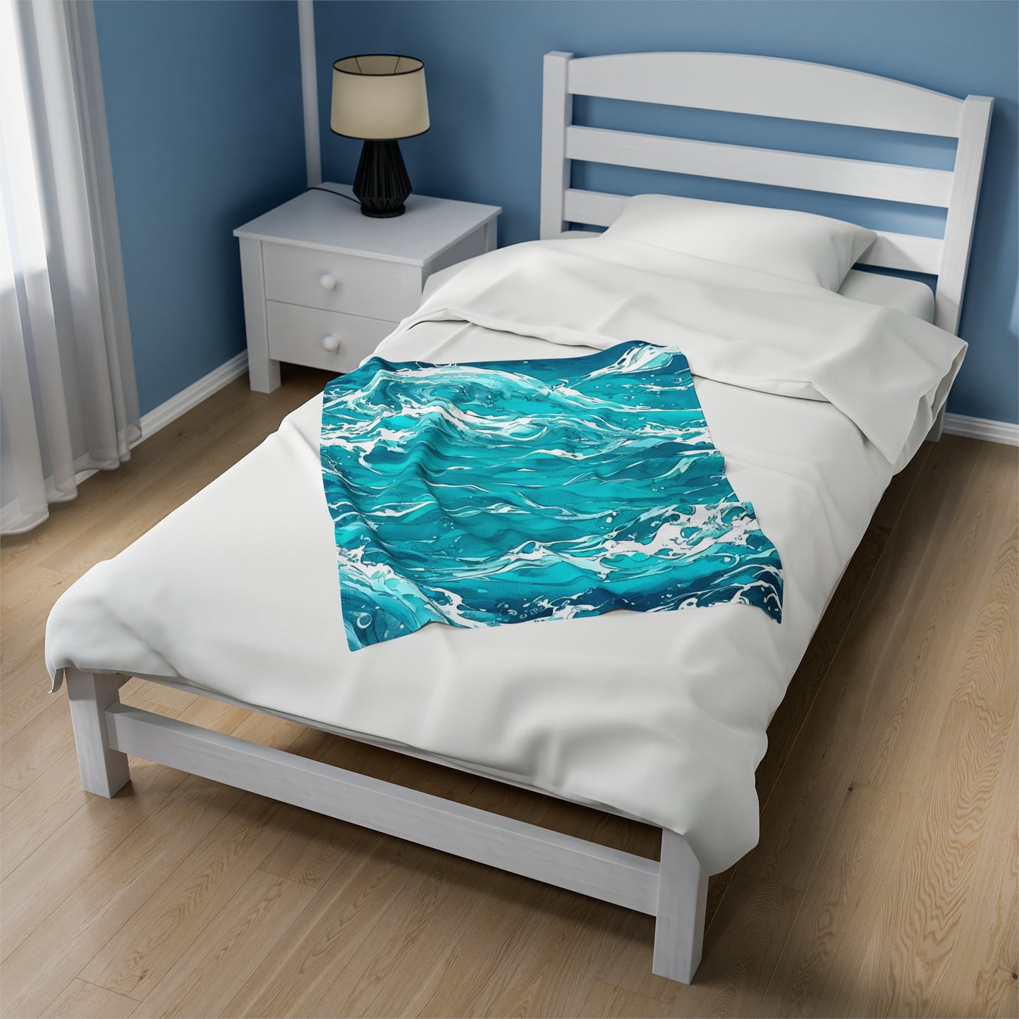 The Swirling Seas blanket design by Lee Hansheng Studios is placed at an angle across a white wooden bed, made with a white duvet, and white wood slat headboard. the blanket design has beautiful waves with mixing hues of blues and greens with foam caps. Light blue walls with white trim, and light wooden floors. A white night stand in distance holds a black lamp with cream lamp shade, Windows on the left have white sheer curtains letting in soft light.