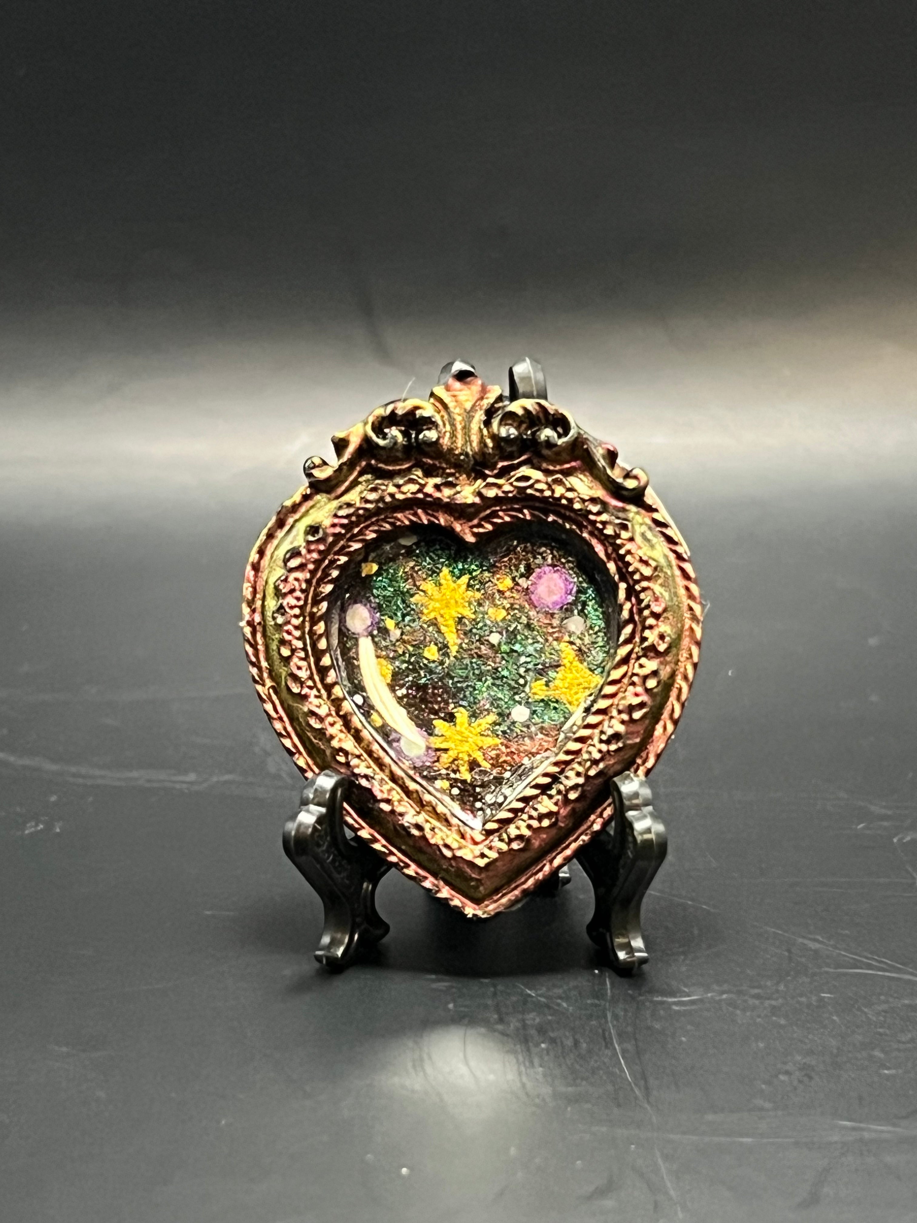 This is the handmade Three Sister Miniature Painting by Chris Foster Design. This is showcasing the front. This is a decorative piece with an heart frame that has been painting a shimmery gold and yellow color, starry night design including yellow stars, sparkling green, orange, yellow and black on a dark background. There are larger yellow stars and smaller white stars. There is a magnet and against a miniature picture frame. 
