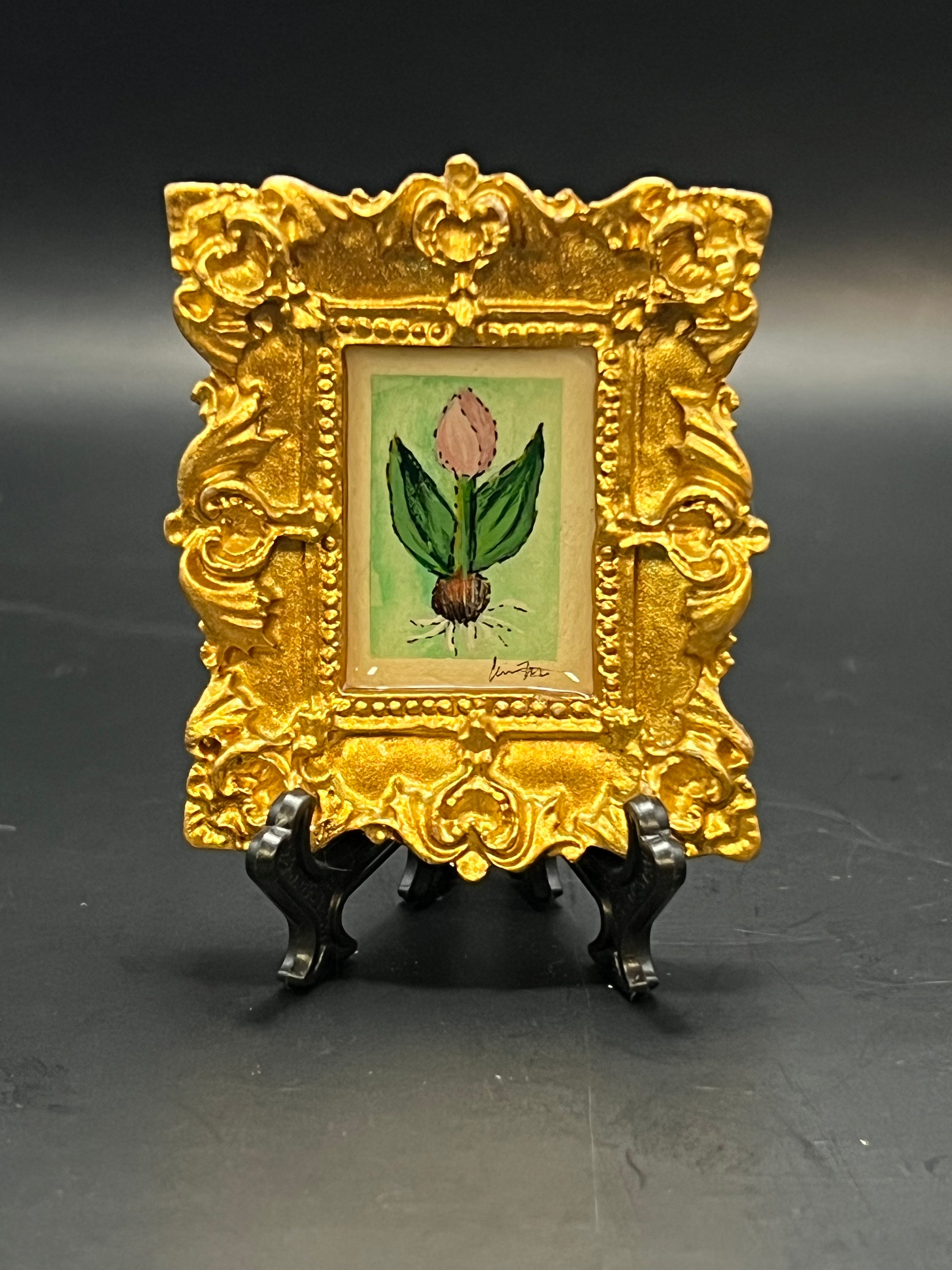 This is the Tulip Mini Painting by Chris Foster Design. This is showcasing the front of the painting. This is a small painting of a pink tulip with two green leaves and the root with the veins sticking out. The painting is on a decorative gold frame on a black stand. It is against a dark background.