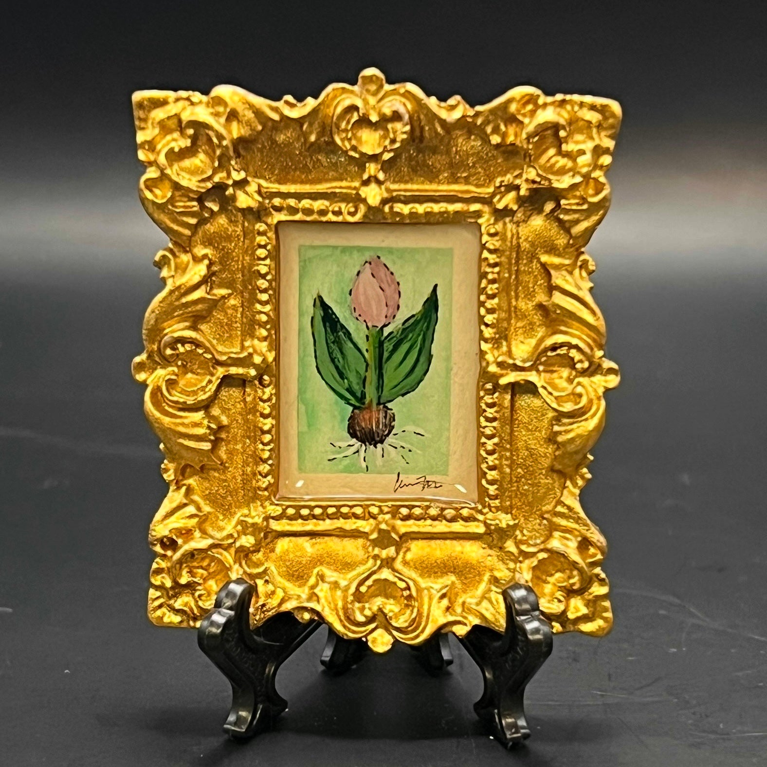 This is the Tulip Mini Painting by Chris Foster Design. This is showcasing the front. Metallic ornate gold frame. Light pink purple tulip on green stem with leaves and brown rooting bulb. Painted on mint green background. It is against a dark background.