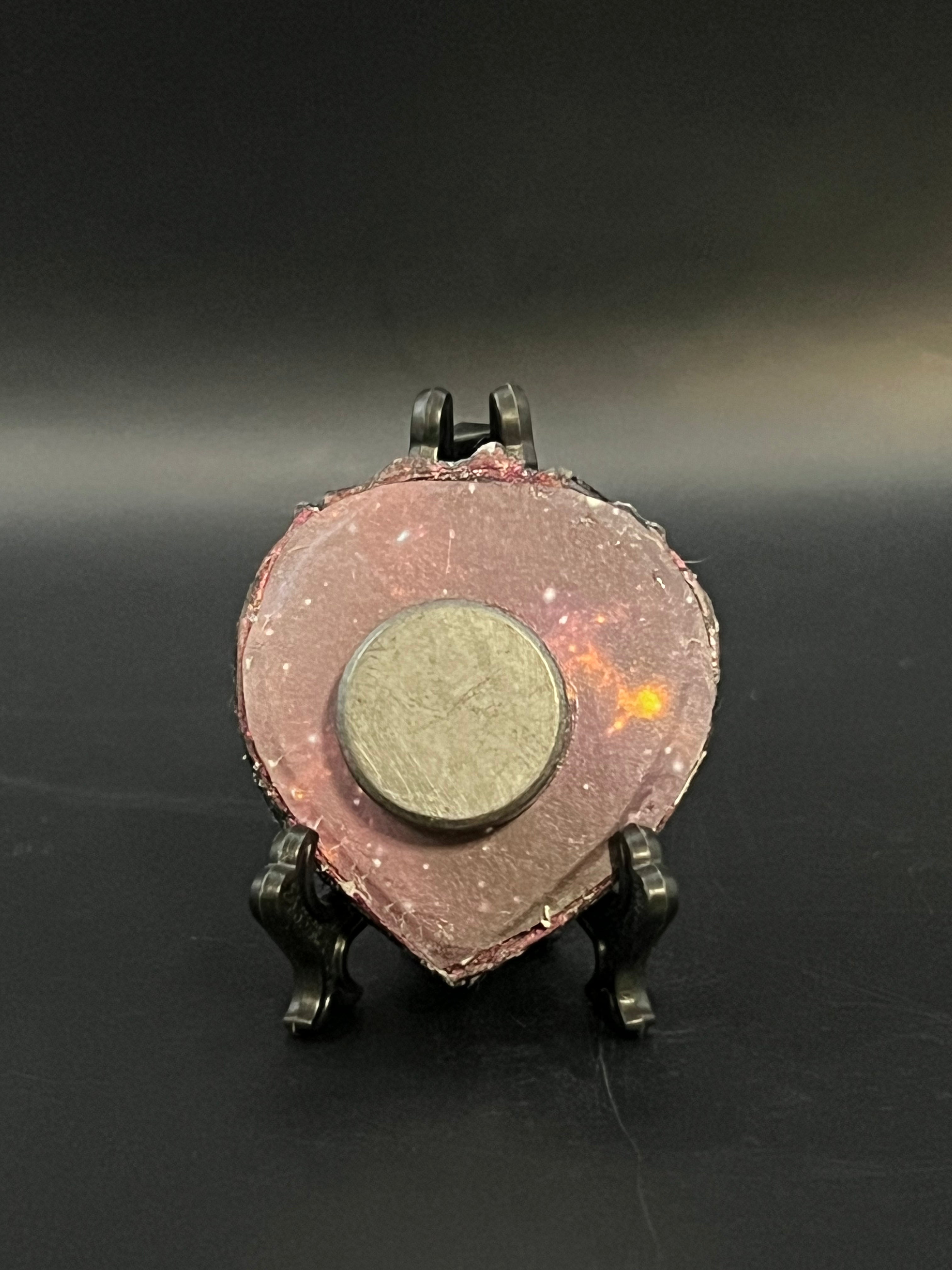 This is the handmade Twinkle in the Void Miniature Painting by Chris Foster Design. This is showcasing the back.  This is a small heart frame and the decorative item with a sparkly mix background with black, orange and little stars on a stand. There is a magnet and against a miniature picture frame.