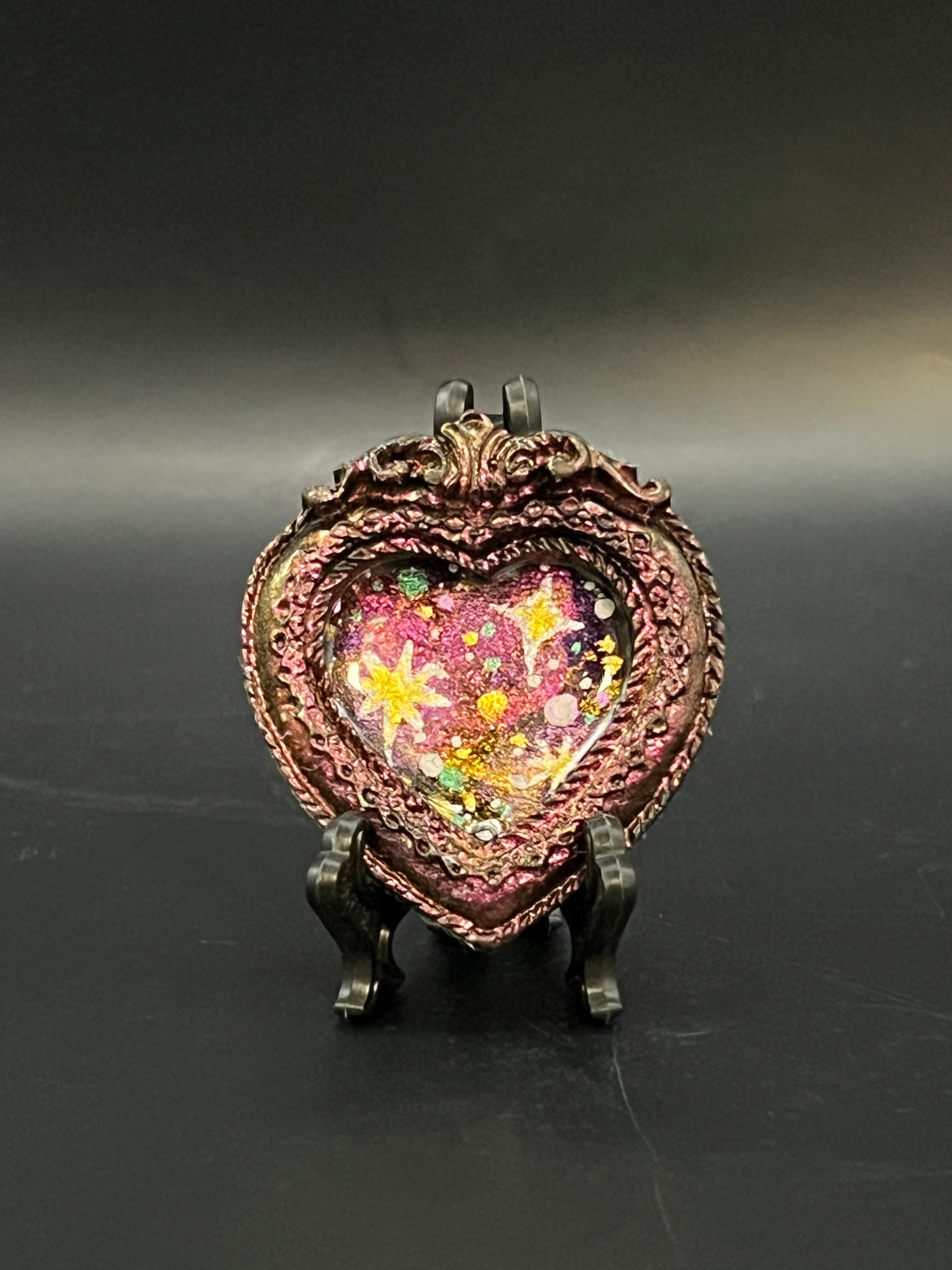 This is the handmade Twinkle in the Void Miniature Painting by Chris Foster Design. This is showcasing the front. This is a decorative piece with a shimmery red heart frame, starry night design including yellow stars, sparkling red mostly with, orange, black, green and white on a dark background. There is a magnet and against a miniature picture frame.