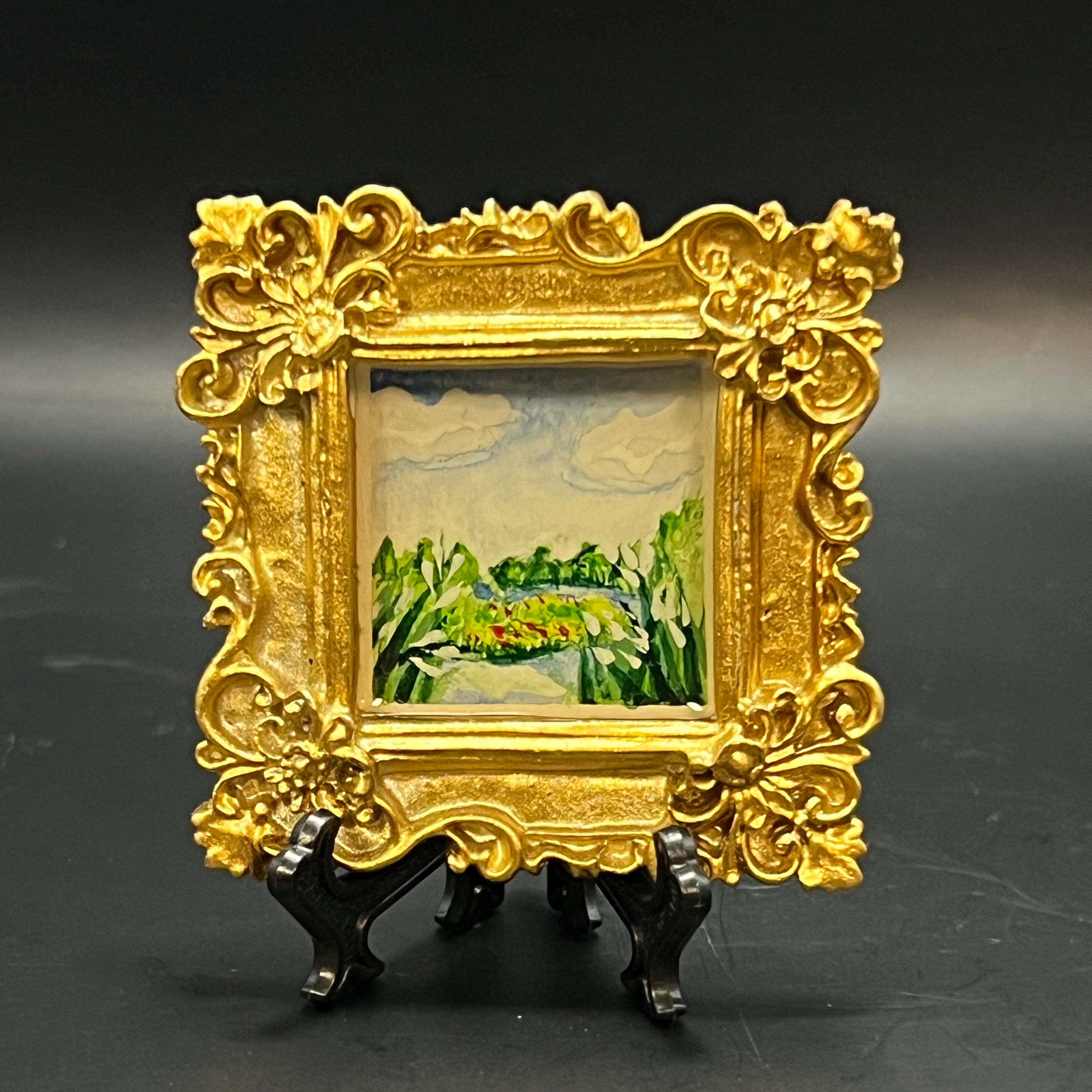 This is the Valley Gold Mini Painting by Chris Foster Design. This is showcasing the front where the ornate gold rectangular frame. The illustration is a landscape river painting with yellow, red, and white flowers and clouds. It is against a dark background. 