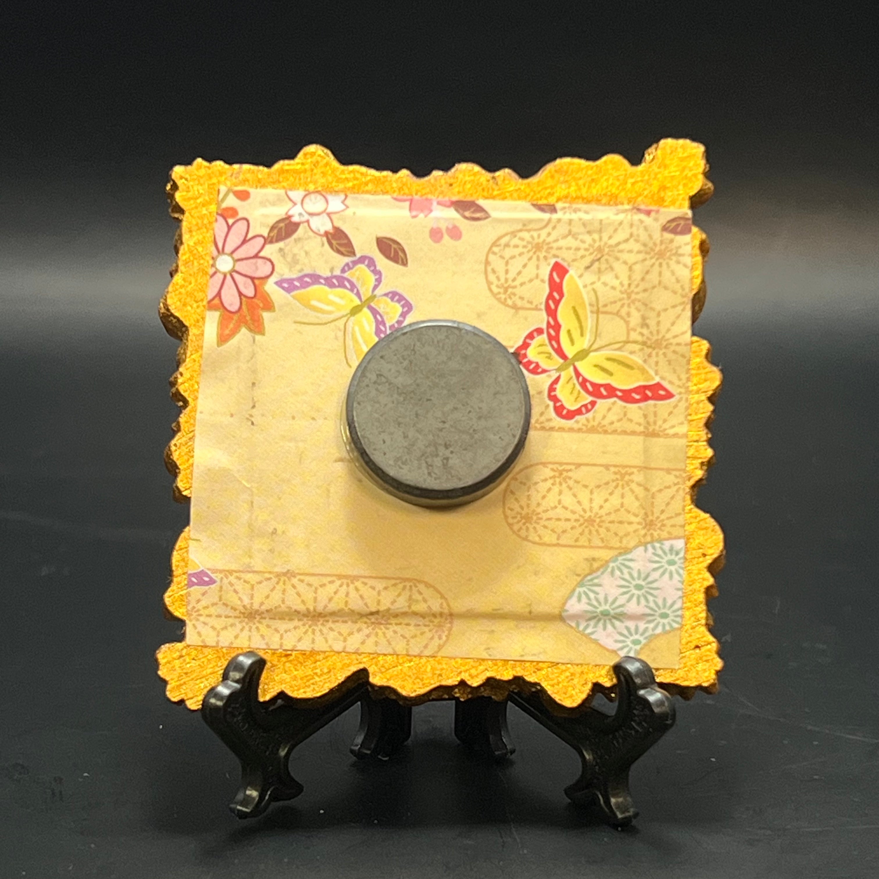 This is the White Perfume Lily by Chris Foster Design. This is showcasing the back. There is a decorative gold frame, black stand, backed with a gold floral design decorated with red/pink flowers, butterflies and orange leave.  A round magnet is on the middle against a dark background.