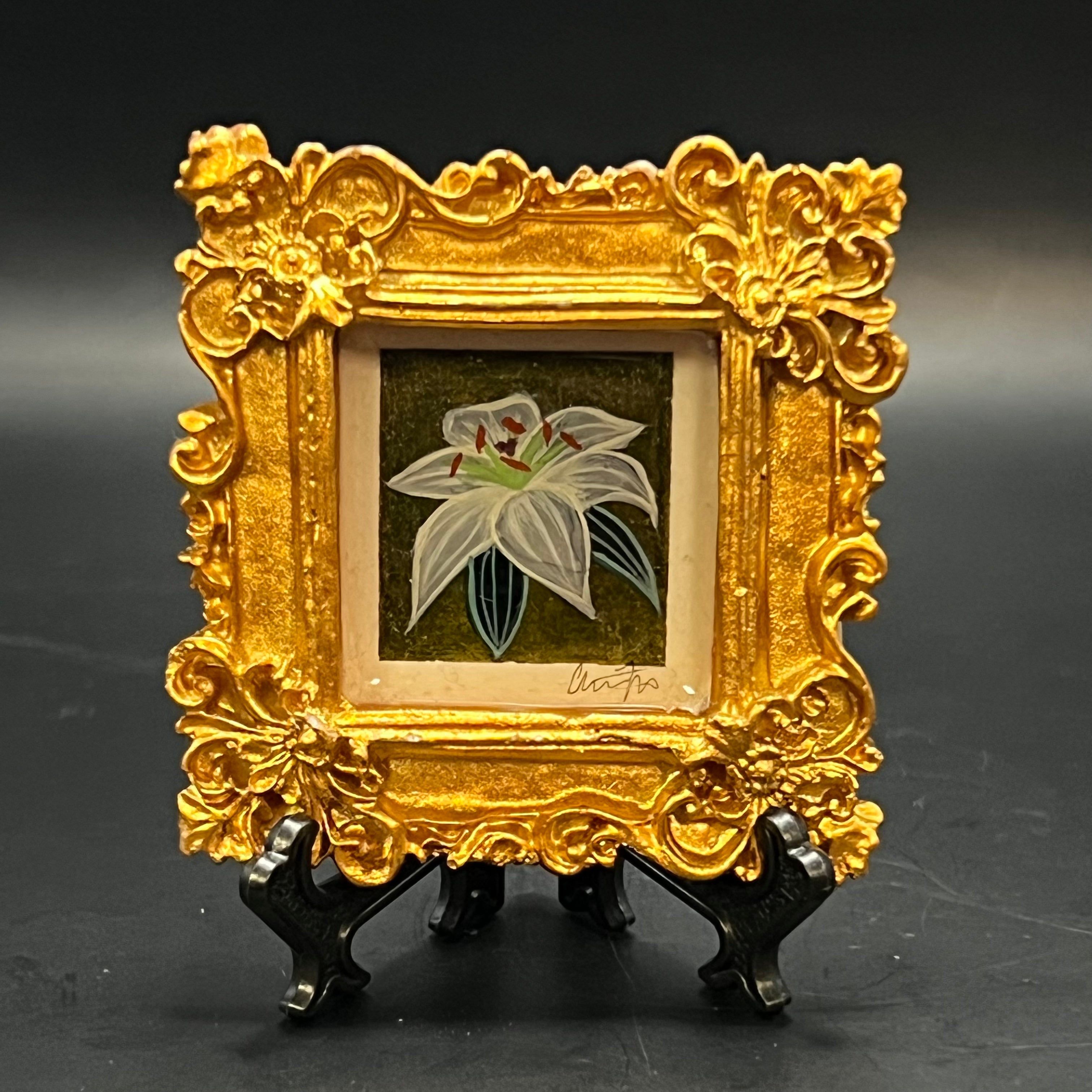 This is the White Perfume Lily Mini Painting by Chris Foster Design. This is showcasing the front. It is a decorative square gold frame with a small painting of the lily which is transluscent and lines of white and green on leaves and flowers respectfully. It is against a dark shimmery gold background. The mini is on a stand and against a dark background.