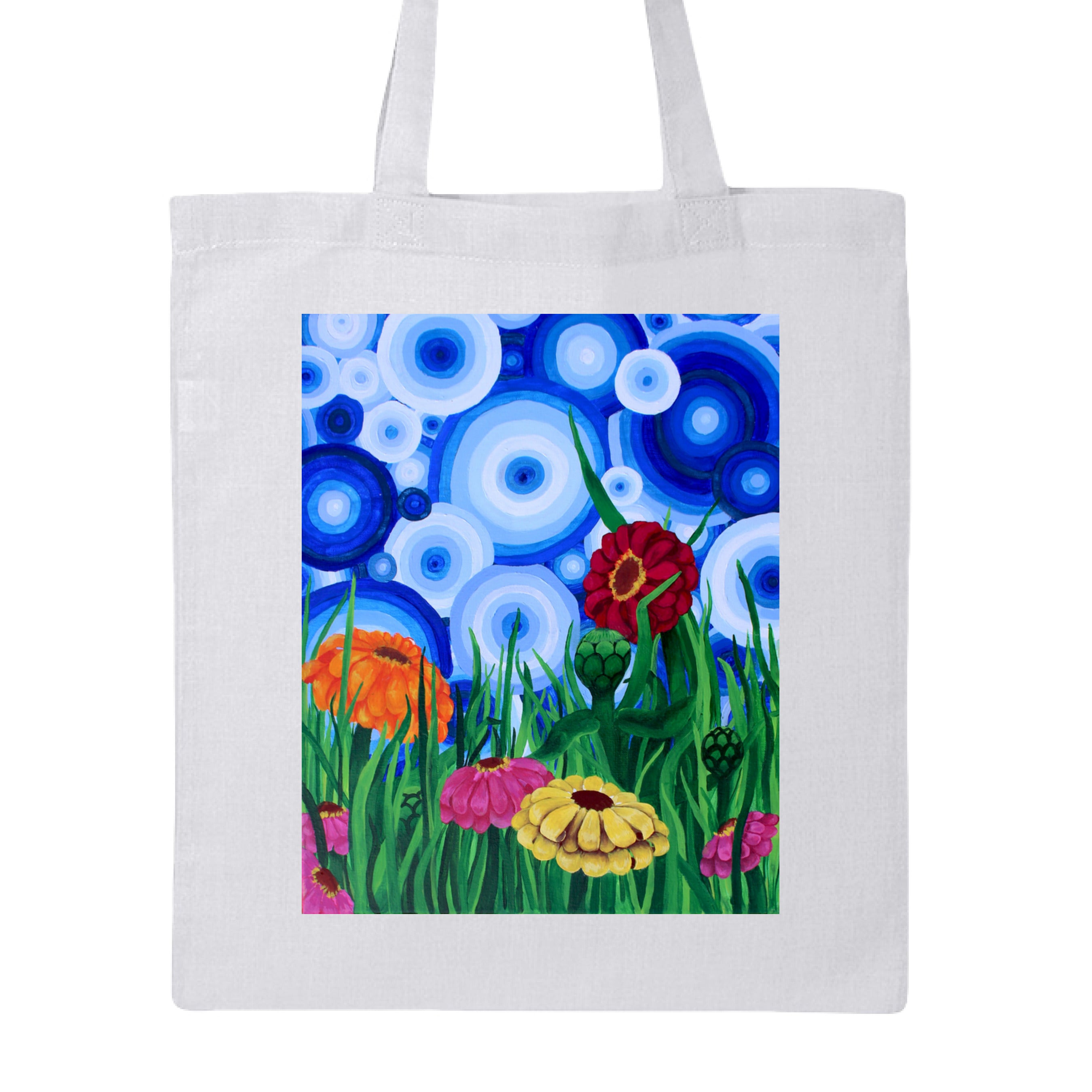 This is the White Zinnia 2 Canvas Tote by Storm Garden Studio. This showcases the front of the tote. The rectangular illustration are bold and color zinnias like red, yellow and orange across tall green grass. The background has many different circles in blue, dark blue and white. The tote is against a white background.