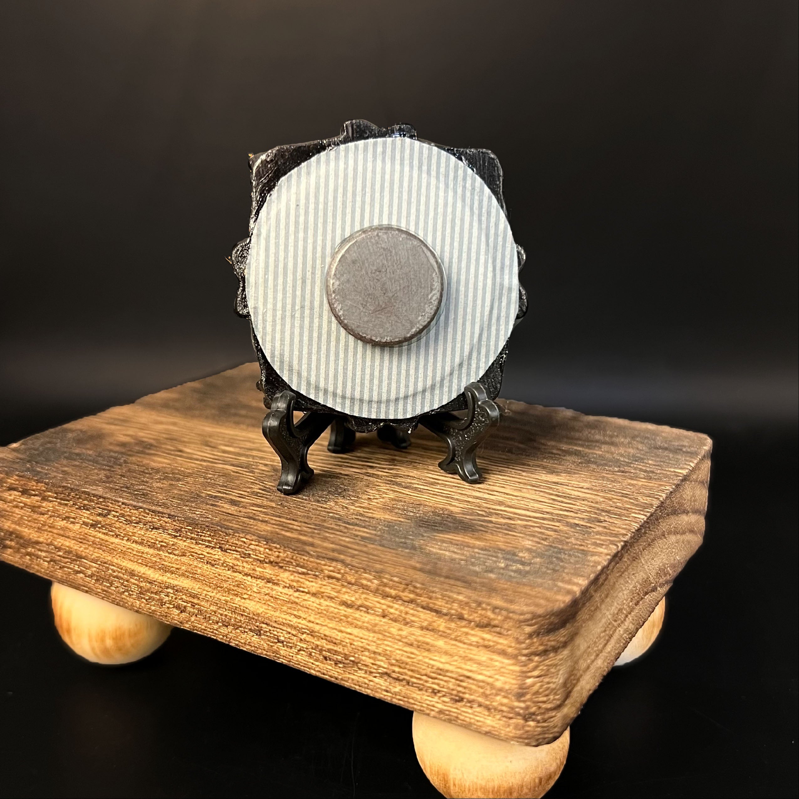 This is the Witch Hat Mini Painting by Chris Foster Design. This is showcasing the back of the painting. The painting has a black circular frame backed with a gray and white background. There is a round circular magnet in the middle. The painting is on a black stand all together on a wooden stand against a dark background.