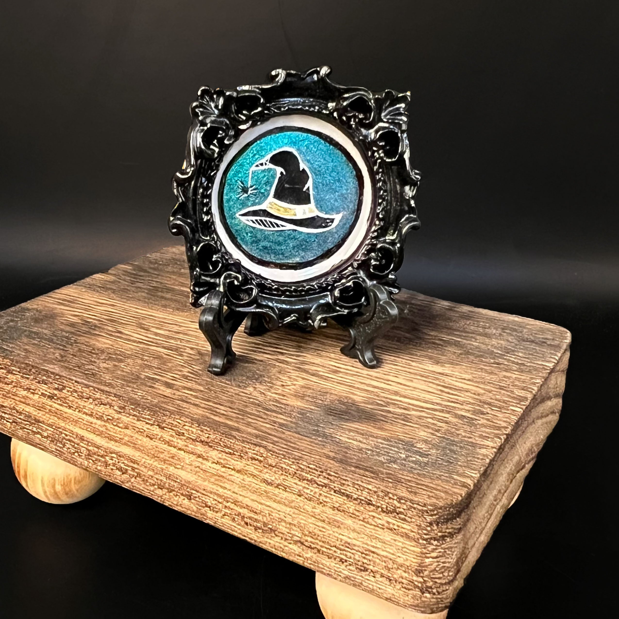 This is the Witch Hat Mini Painting by Chris Foster Design. This is showcasing the front of the painting. The painting has a black circular frame with a black hat, the tip of the hat has a spider hanging, a yellow strip at the base of the hat, outlined in white on a shimmery light blue background. The painting is on a black stand all together on a wooden stand against a dark background.